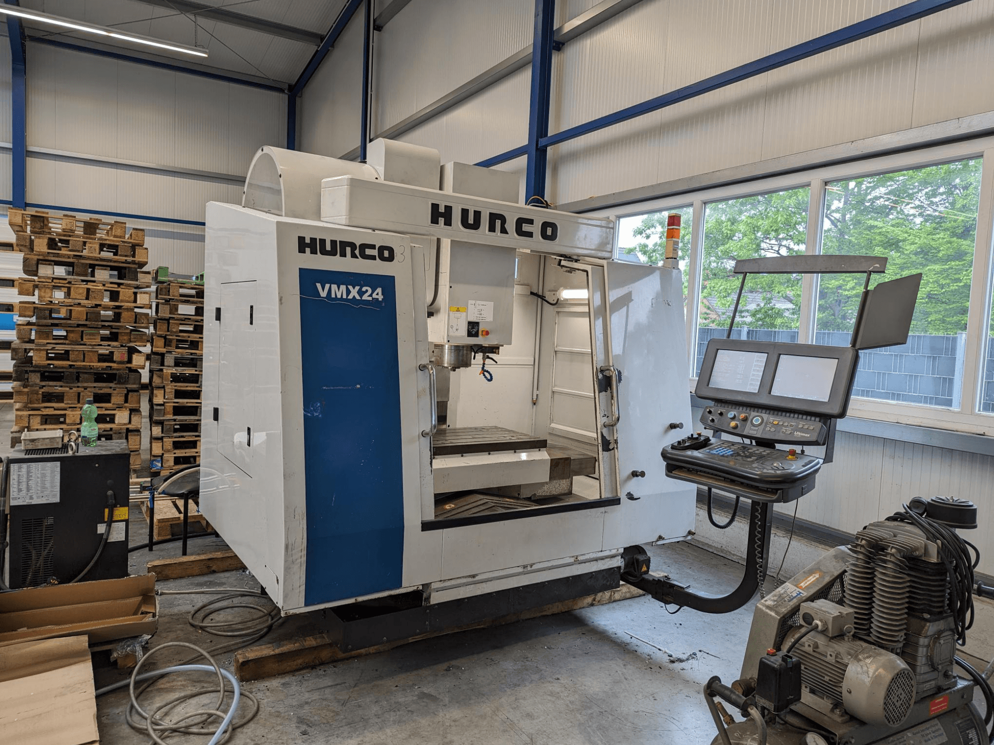 Hurco VMX24 fräsmaskin, framifrån, med kontrollpanel och verktygsområde; staplade pallar i bakgrunden; industriell arbetsplatsmiljö.
