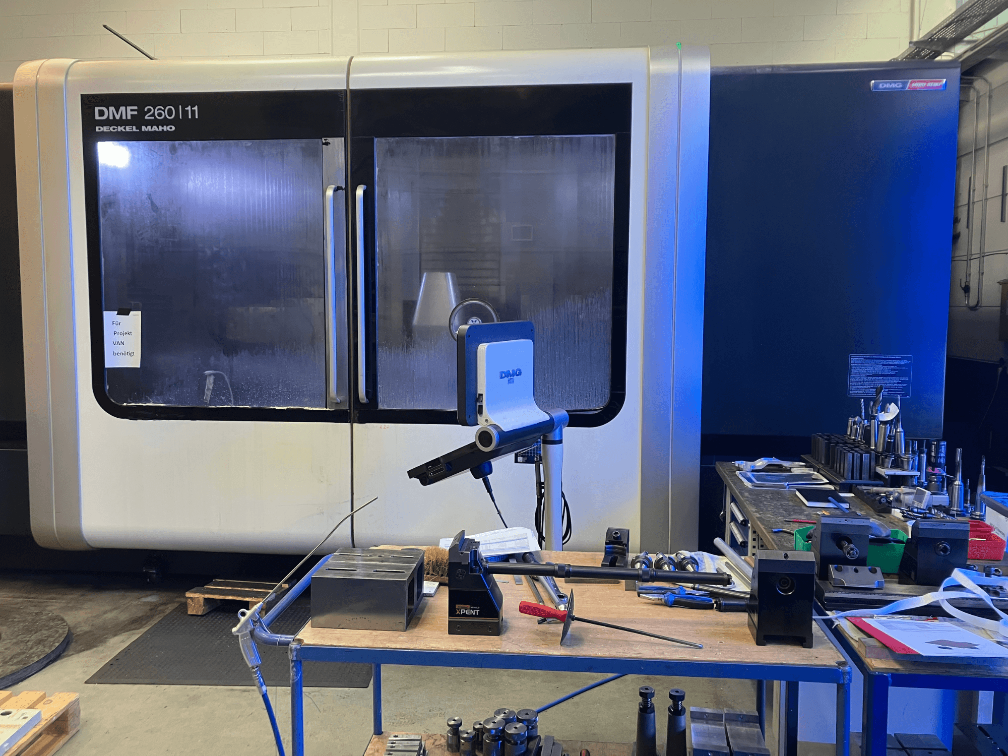 Frontvy av DMG DECKEL MAHO DMF 260|11 vertikal bearbetningscenter med verktyg och material på en arbetsstation.