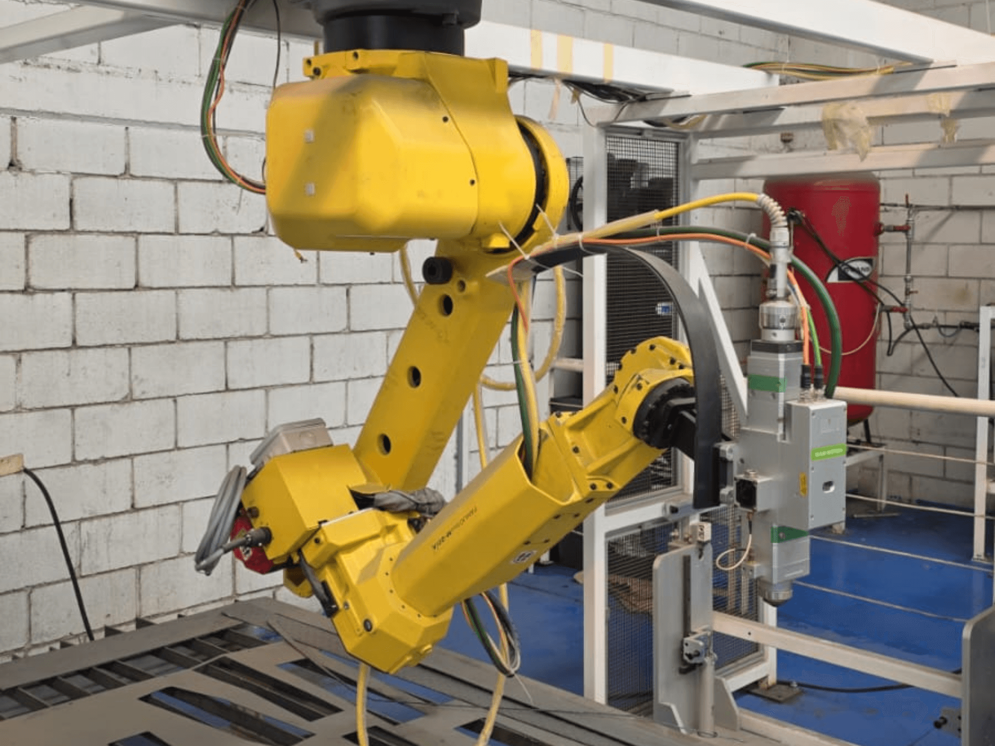 Gul robotarm med ett precisionsverktyg, med flera leder och kablar, sedd framifrån i en industrimiljö.