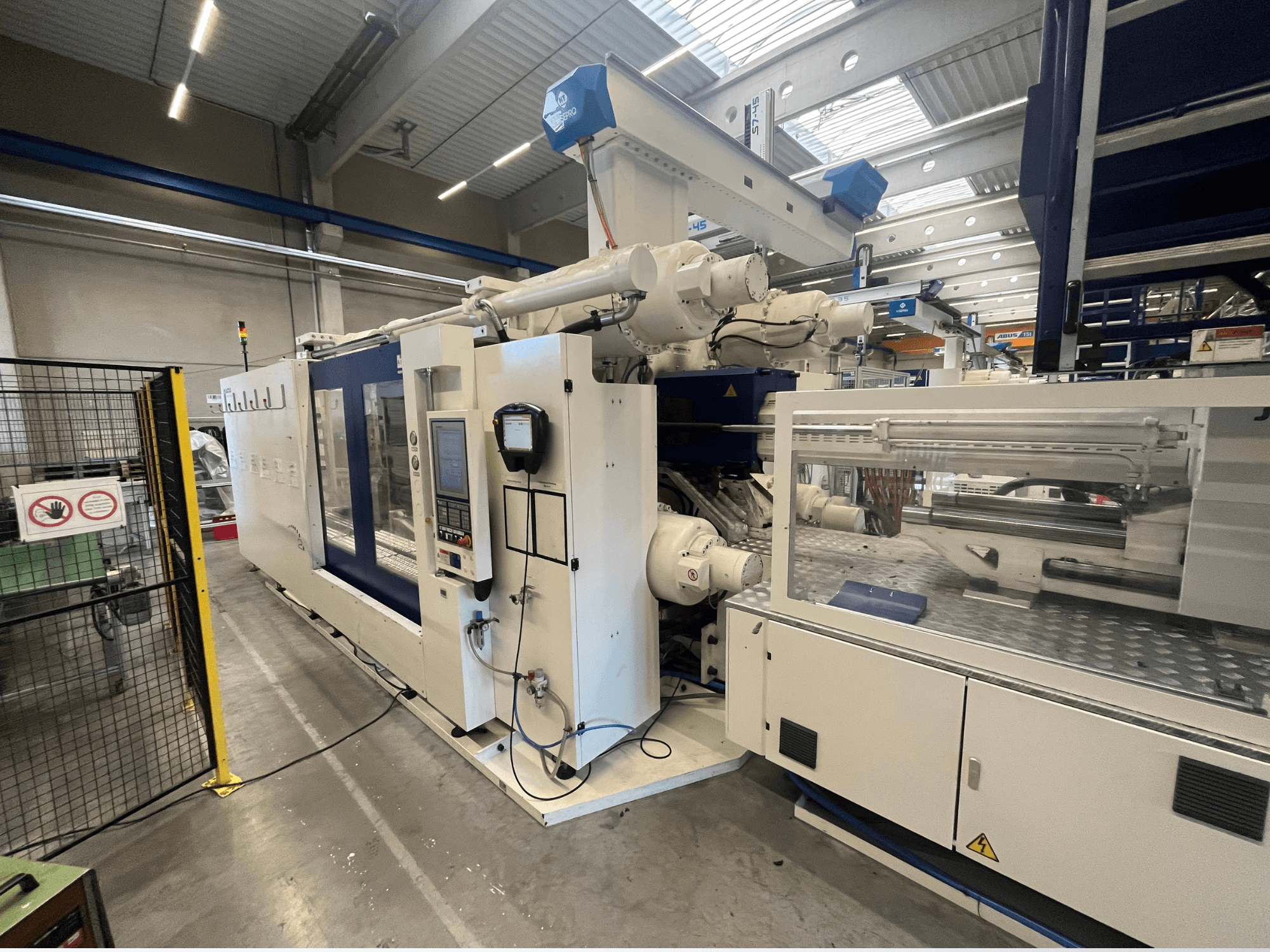 Formsprutningsmaskin i vitt och blått, framifrån, med kontrollpanel, robotarm och säkerhetsskyltar.