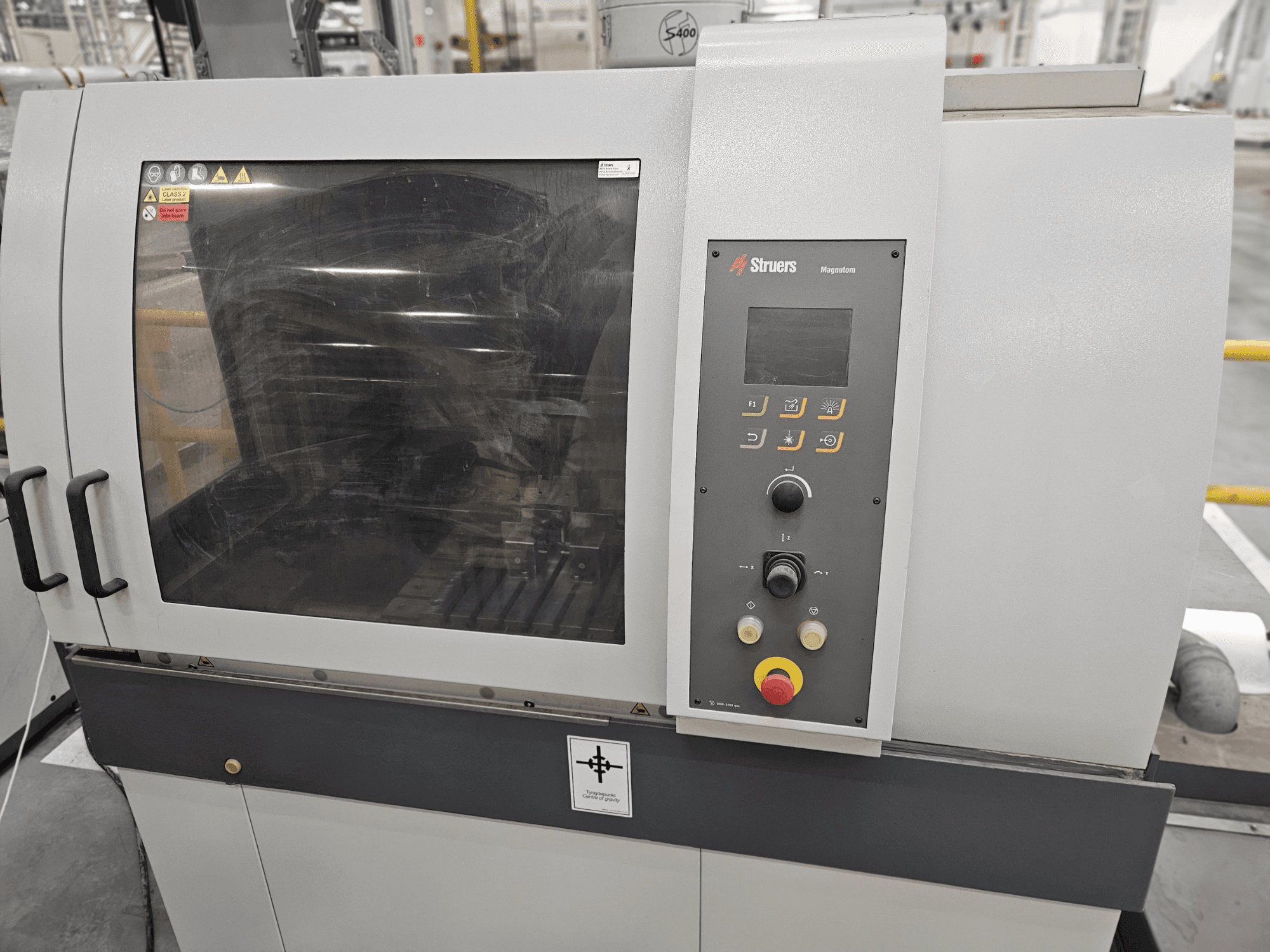 Struers Magpentor maskin framifrån med kontrollpanel, knappar och ett stort visningsfönster. Tekniska etiketter synliga.