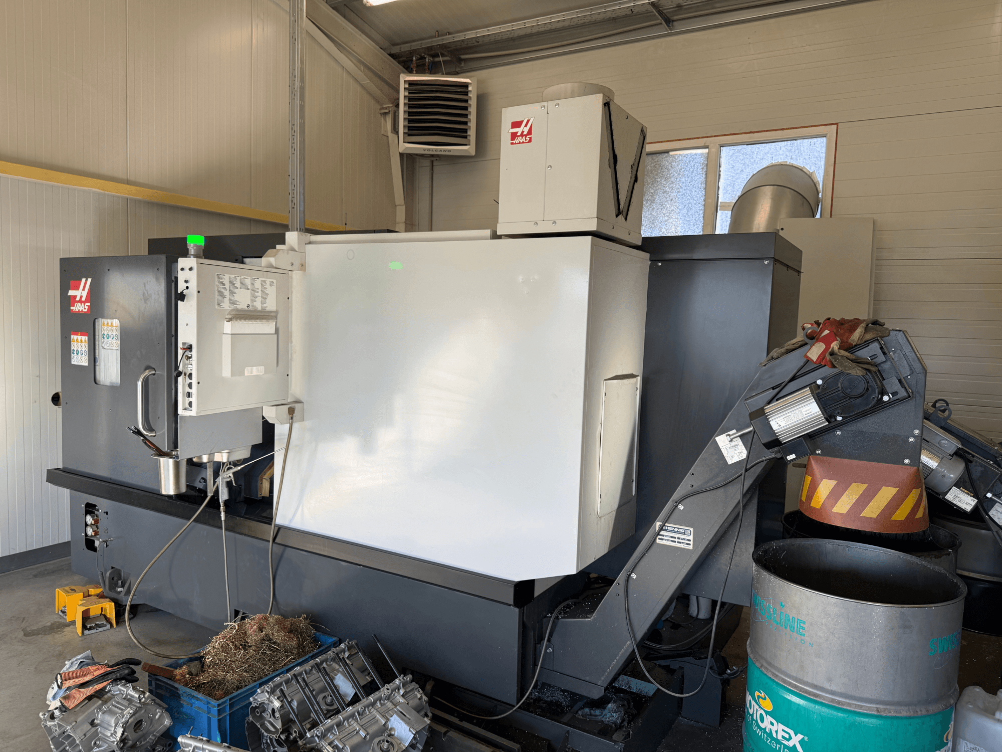 Haas CNC-maskin med kontrollpanel, vy från sidan; synliga kyl- och oljeledningar; metallskrot i blå behållare; industrimiljö.