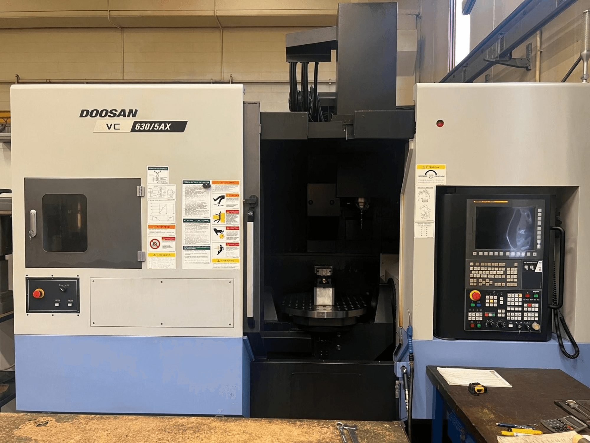 Framifrånvy av ett vertikalt bearbetningscenter av typen DOOSAN VC 630/5AX, där kontrollpanelen och spindeldelen syns tydligt med synliga säkerhetsetiketter.