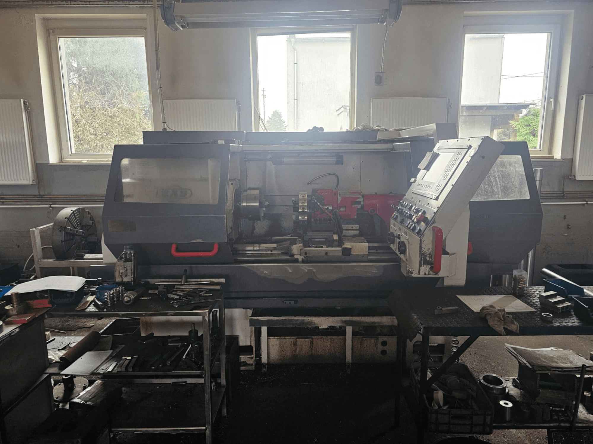 Svarv i en verkstad, framifrån, med kontrollpanel och verktyg på bord, omgiven av fönster och rörigt arbetsutrymme.