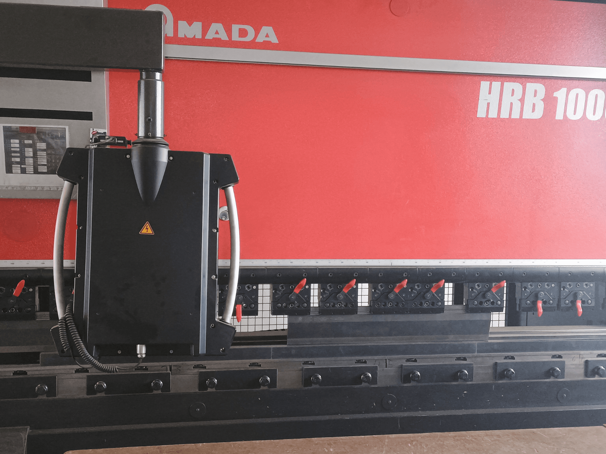 Amada HRB 100 bockningsmaskin framifrån; har ett svart styrhuvud med ett koniskt munstycke och justerbara klämmor.
