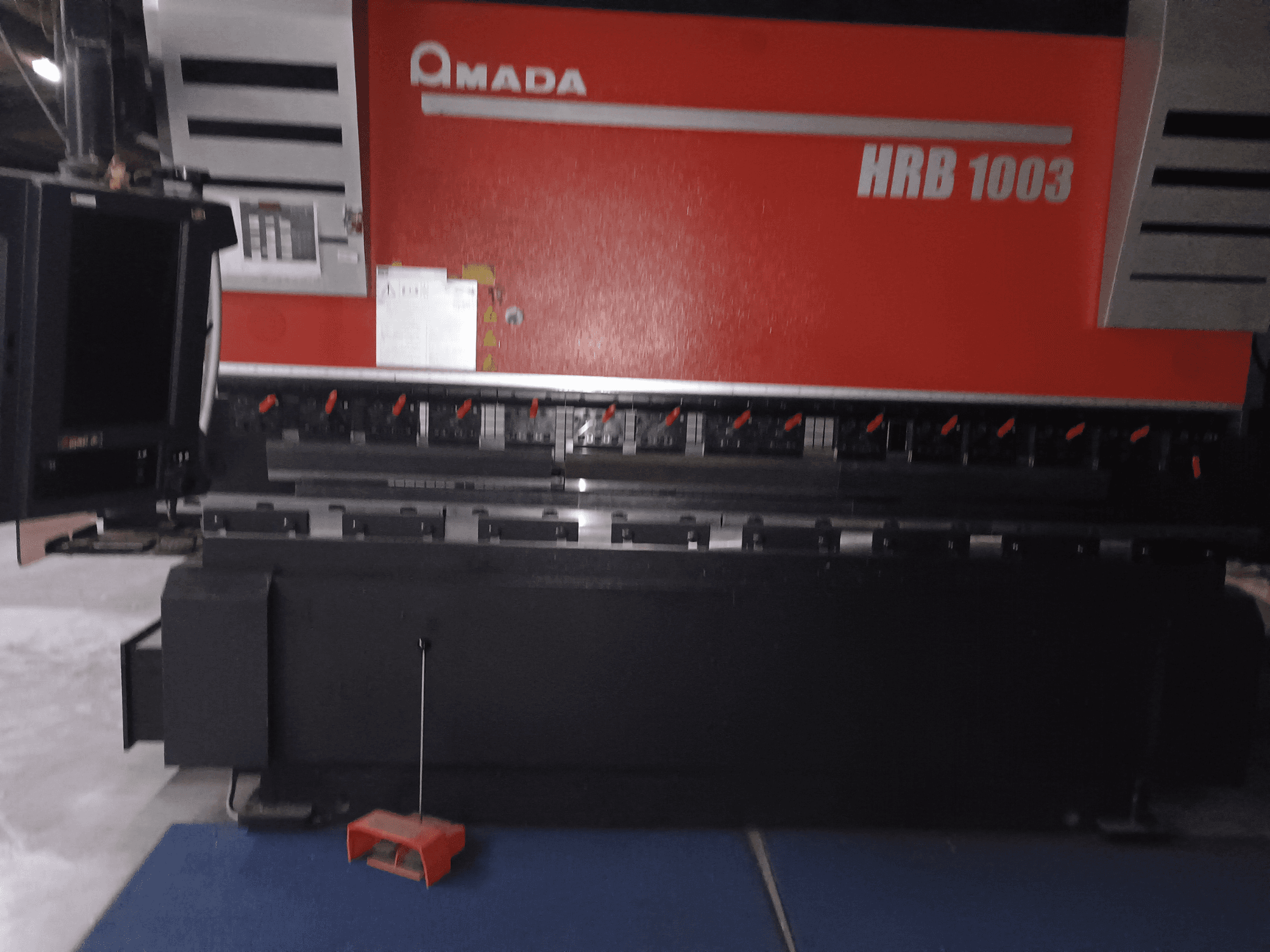 Amada HRB 1003 kantpress, framifrån; har kontrollpanel med röda knappar och svart hölje, placerad i en industriell miljö.