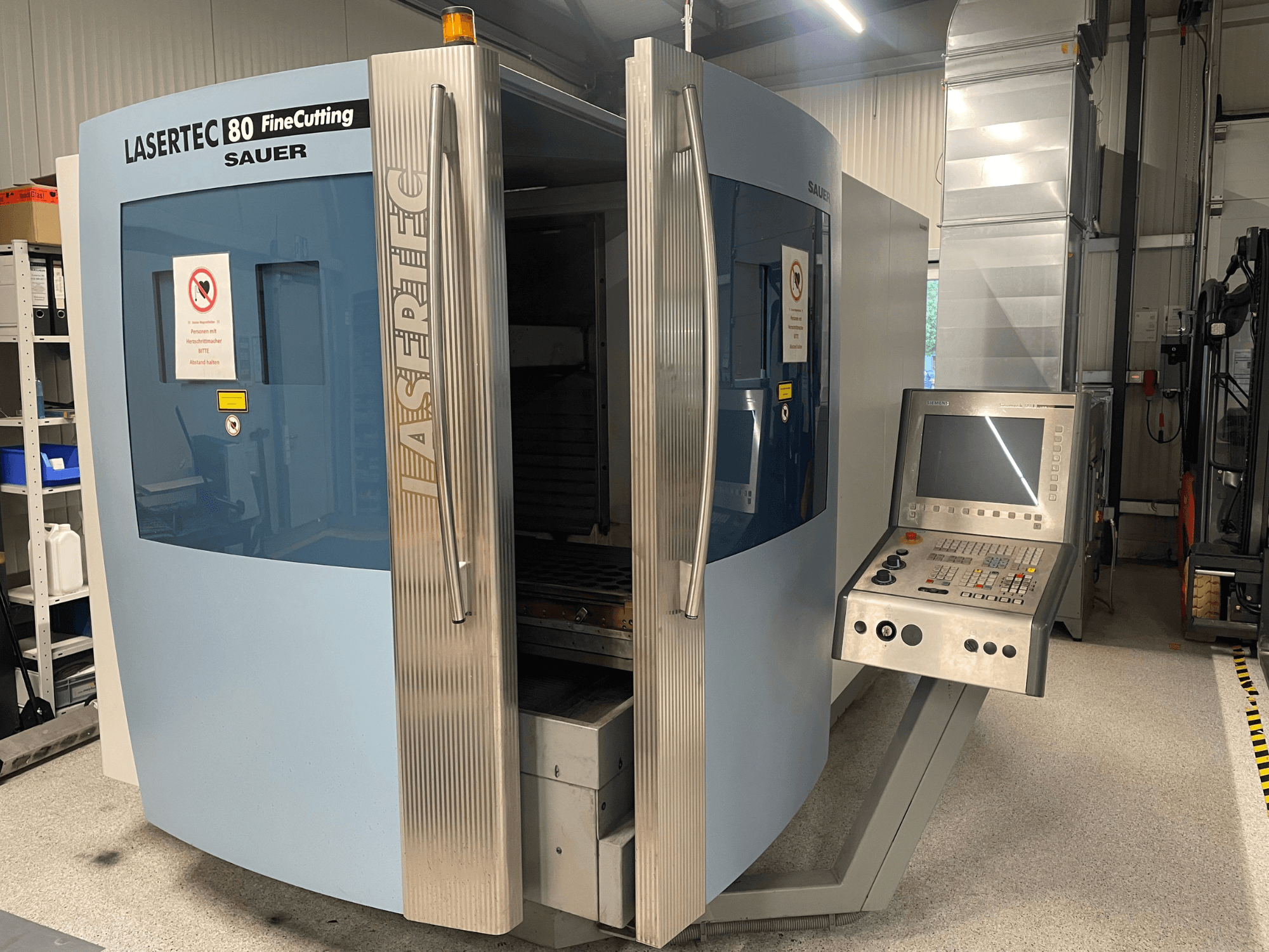 Framifrånvy av DMG Sauer Lasertec80 FineCutting-plåtbearbetningsmaskin med kontrollpanel, öppna luckor och synliga säkerhetsskyltar.