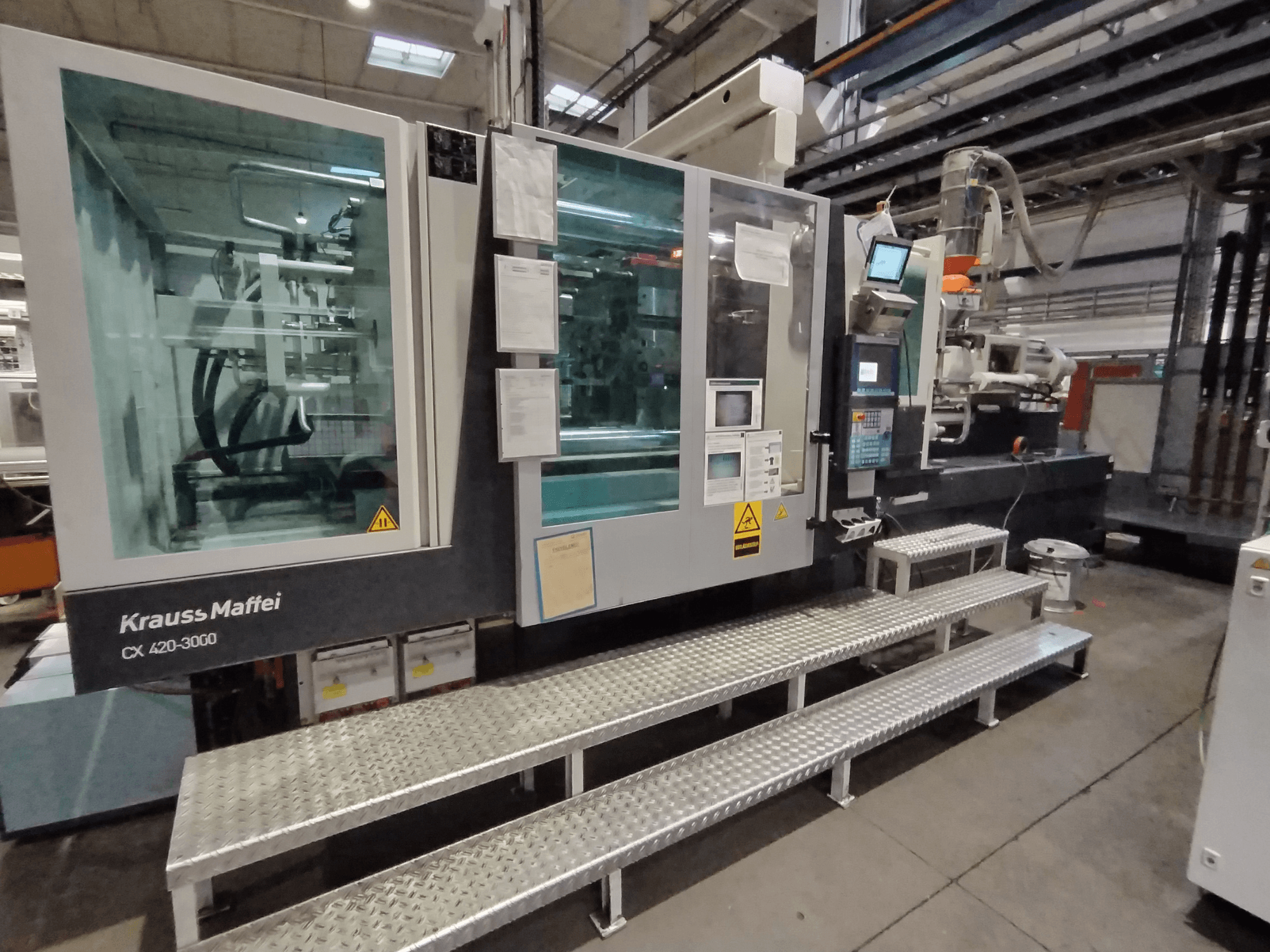 Krauss Maffei CX 420-3000 formsprutningsmaskin, vy från sidan, med synlig kontrollpanel och säkerhetsskyltar, uppställd på metallsteg.