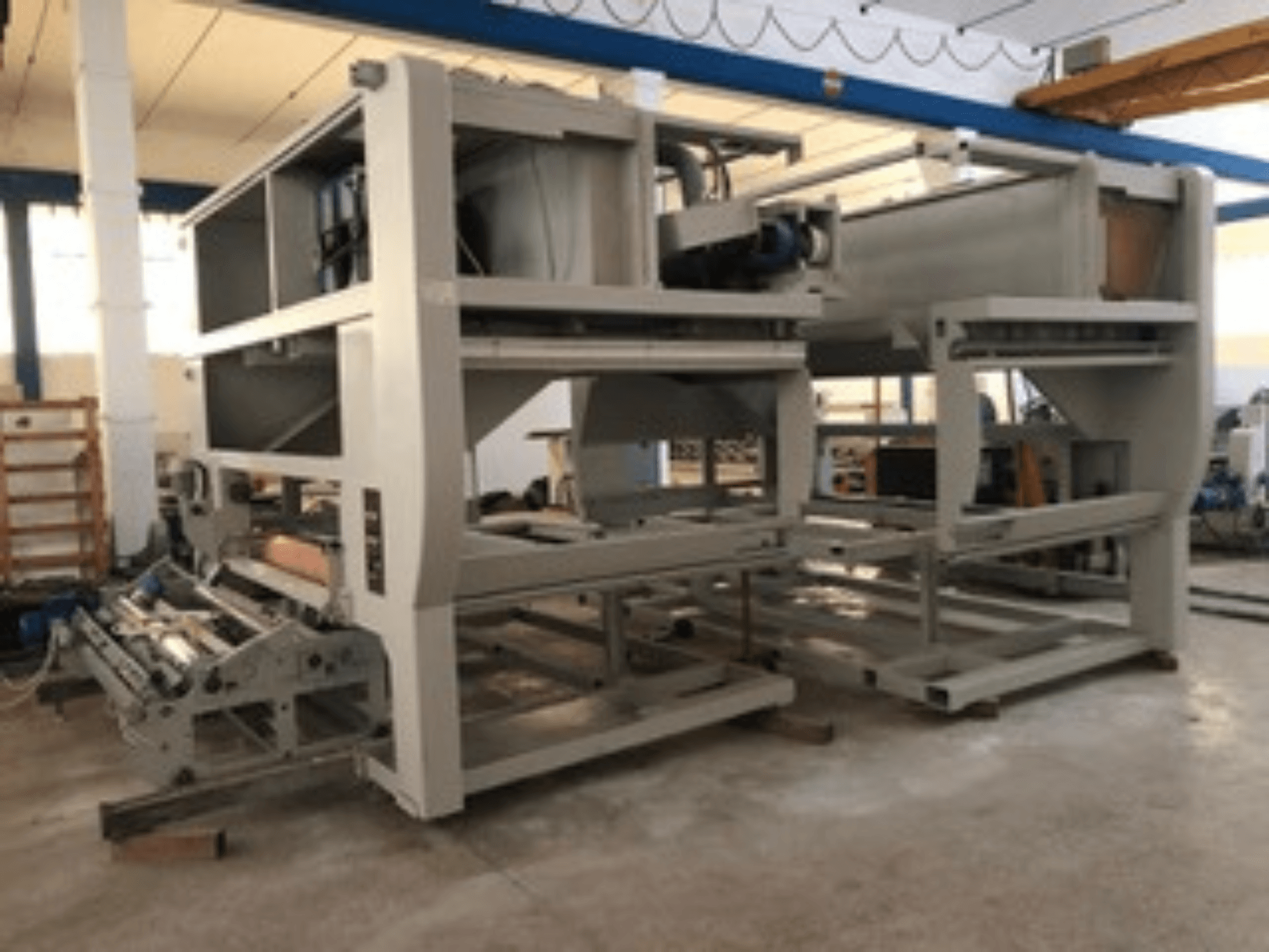 Industrimaskin med flera transportrullar och stor struktur; sidovy, grå färg, placerad i en väl upplyst fabrik.