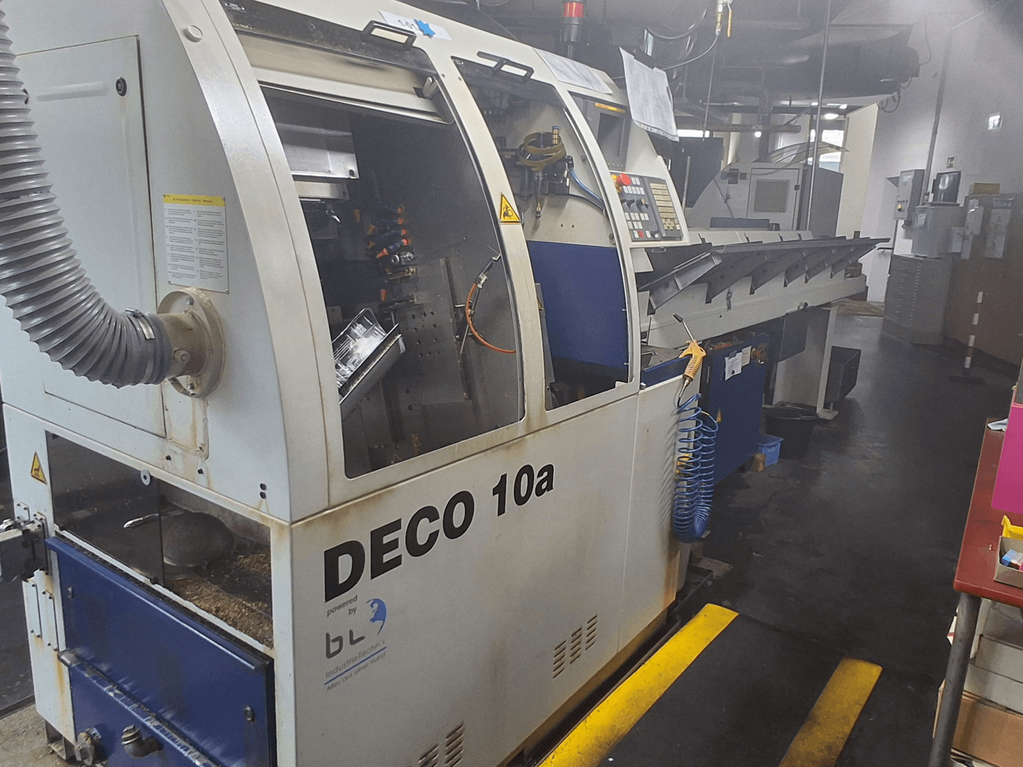 Svarvmaskin av schweizisk typ TORNOS DECO 10a, framifrån; har en kontrollpanel, synliga mekanismer och ett dammutsugningsrör.