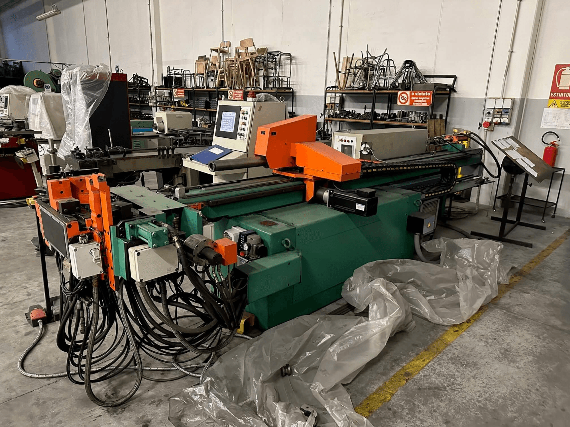 Industrimaskin med orange och grön kaross, kontrollpanel och många kablar, sedd framifrån i en verkstadsmiljö.