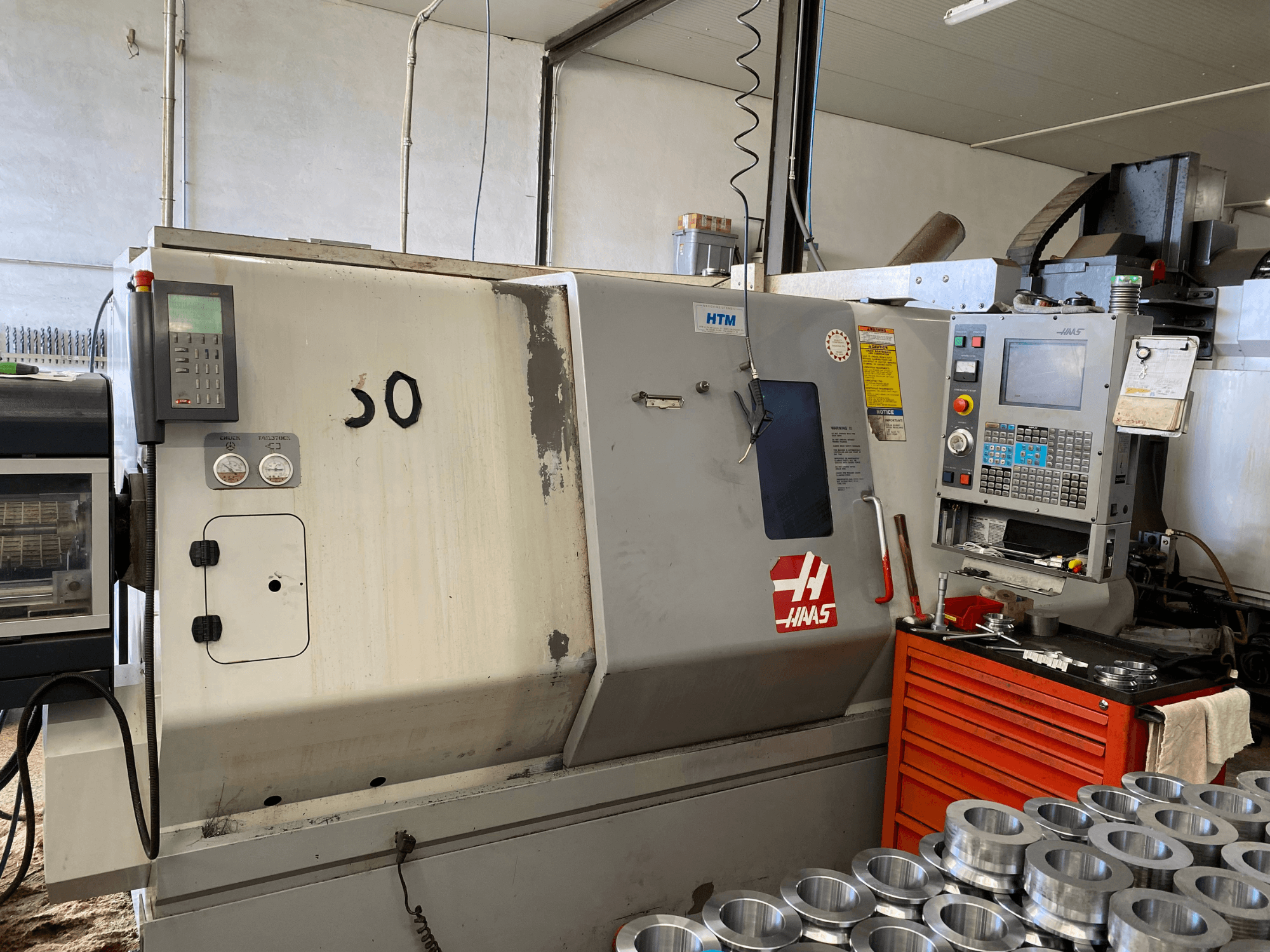 Haas CNC-maskin med kontrollpanel, vy framifrån, med manöverknappar, mätare och metalldelar staplade i närheten.