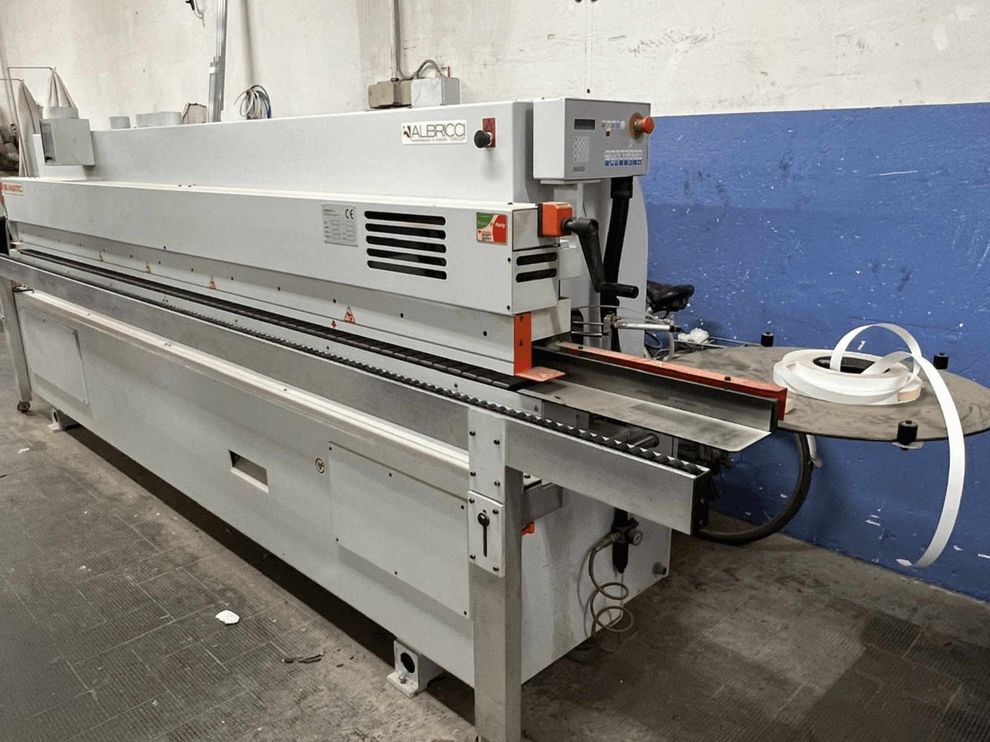 ALBRICO kantlistningsmaskin framifrån, med ett långt transportband och en påmonterad rulle med självhäftande tejp.