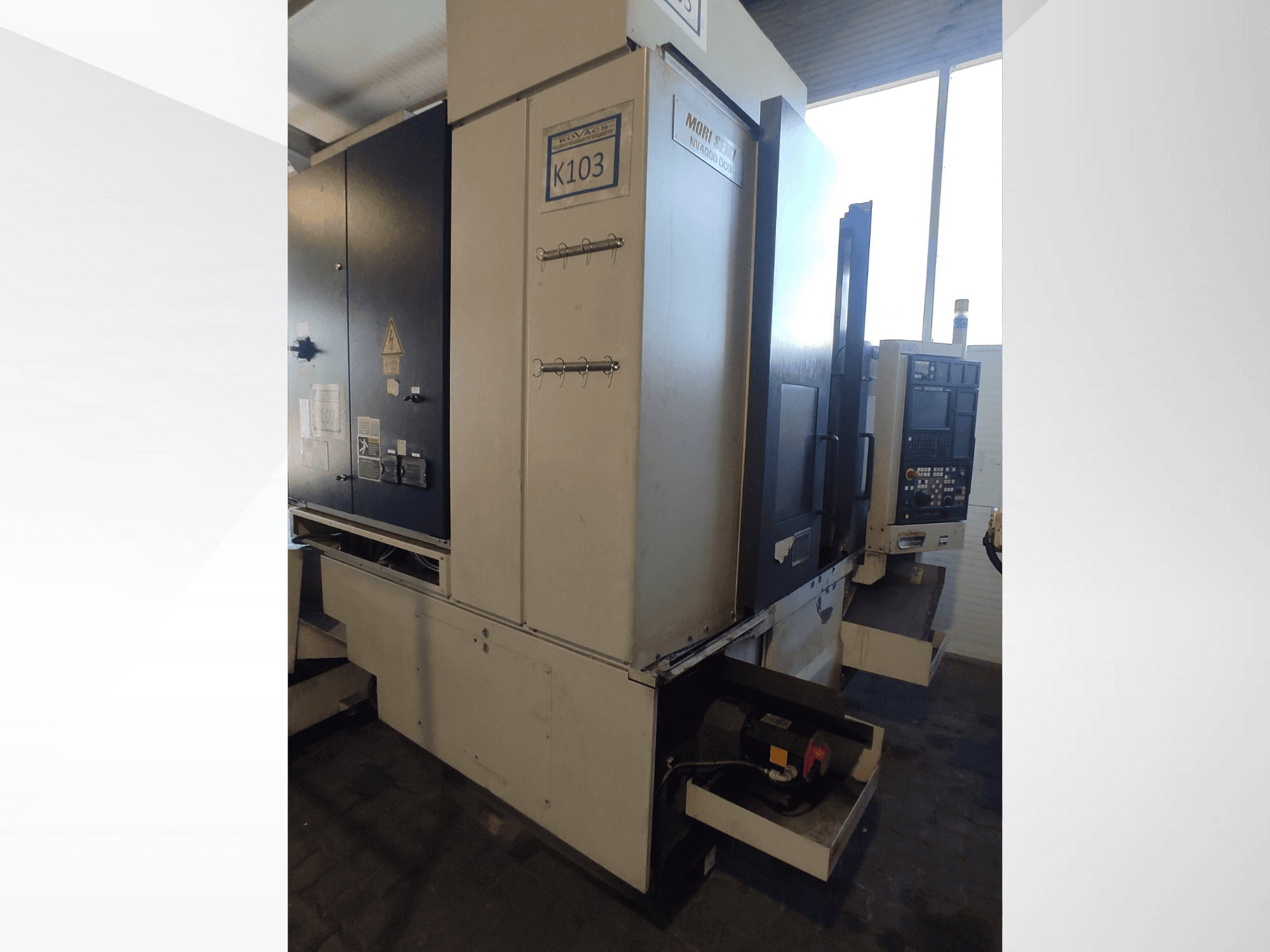 Mori Seiki K103 CNC-maskin framifrån, med kontrollpanel, säkerhetsetiketter och elektriska komponenter synliga.