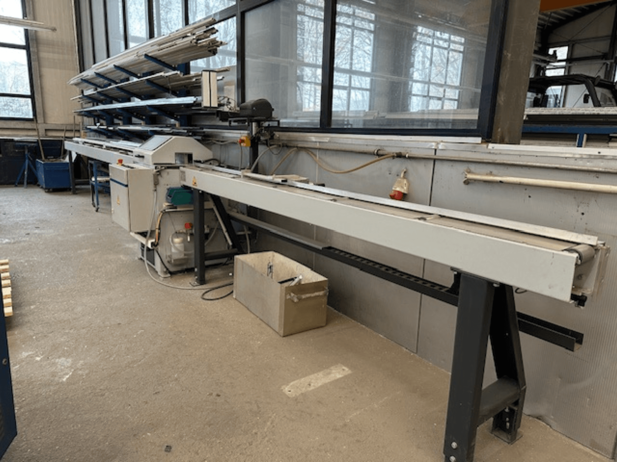 SCHÜCO PGS metallcirkelsåg i en verkstad, sidovy med en justerbar kapbädd och en hylla med metallprofiler ovanför.