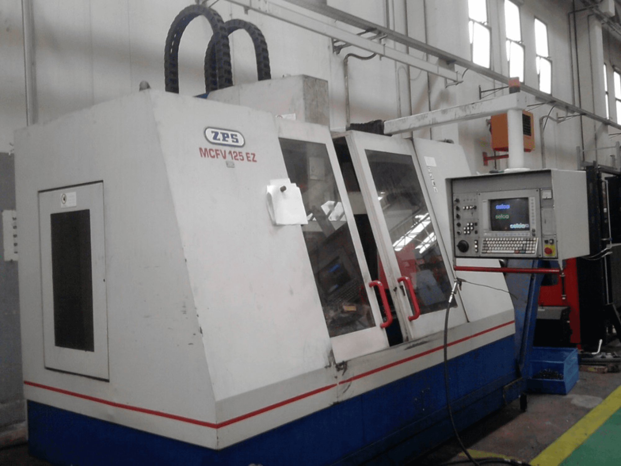 ZPS MCFV 125 EZ CNC-maskin, framifrån med kontrollpanel och röda handtag, i verkstadsmiljö med synliga slangar.
