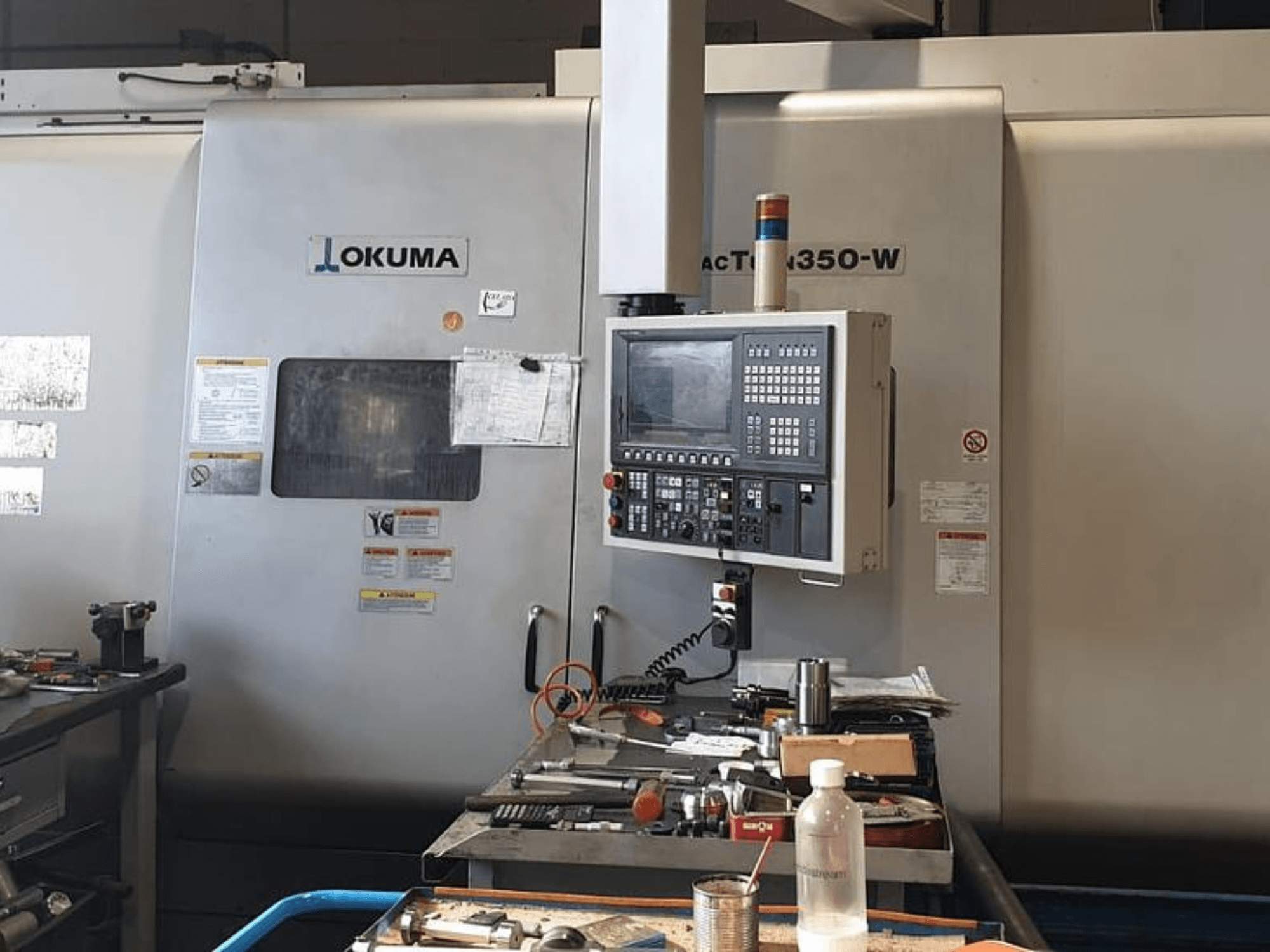 Okuma MAC TURN 350-W svarvfräscenter, framifrån med kontrollpanel och arbetsyta belamrad med verktyg och en flaska.