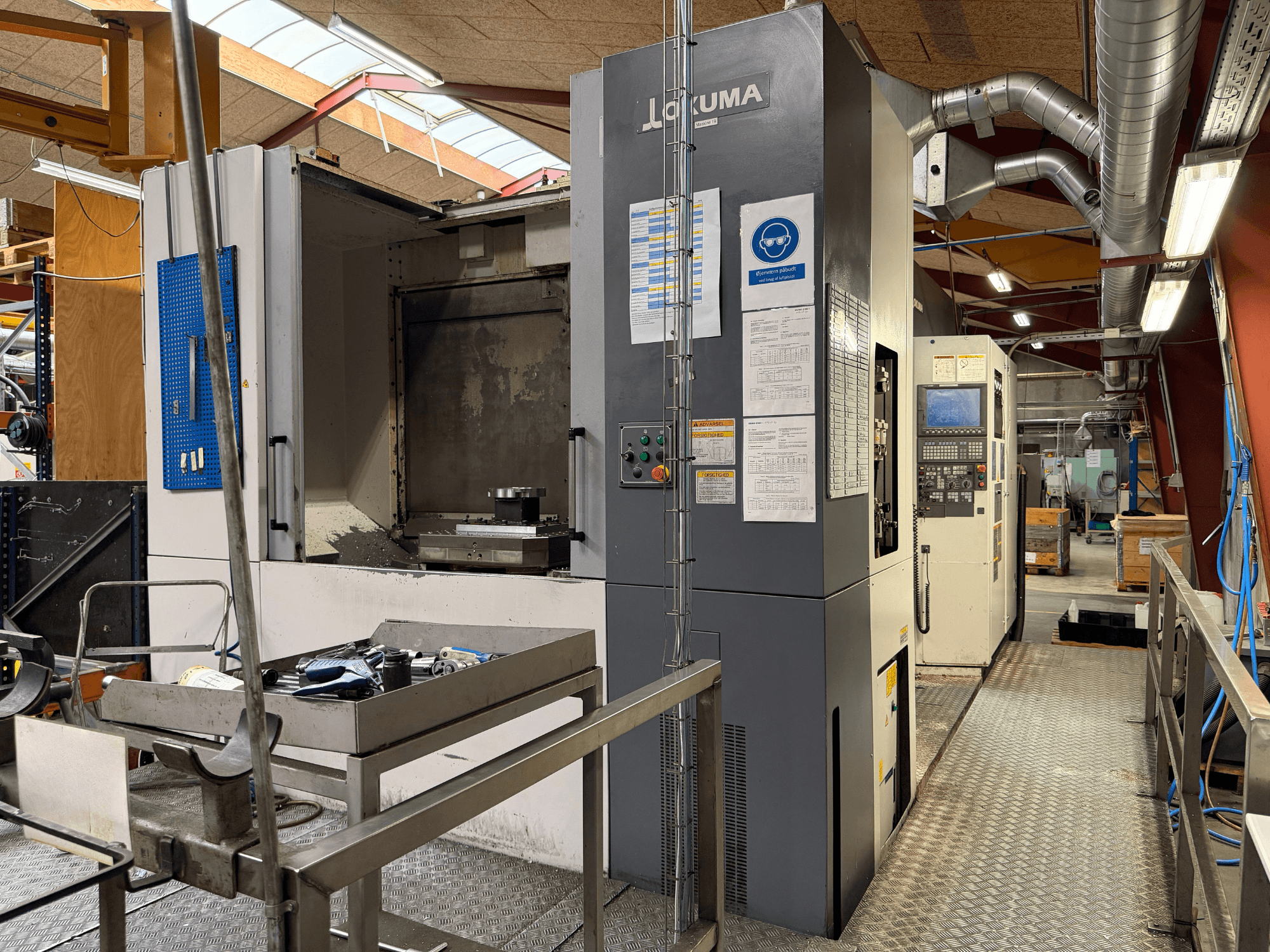 Okuma MA-600HB horisontellt bearbetningscenter, framifrån, med verktyg på ett metallbord och maskinens manöverpaneler.
