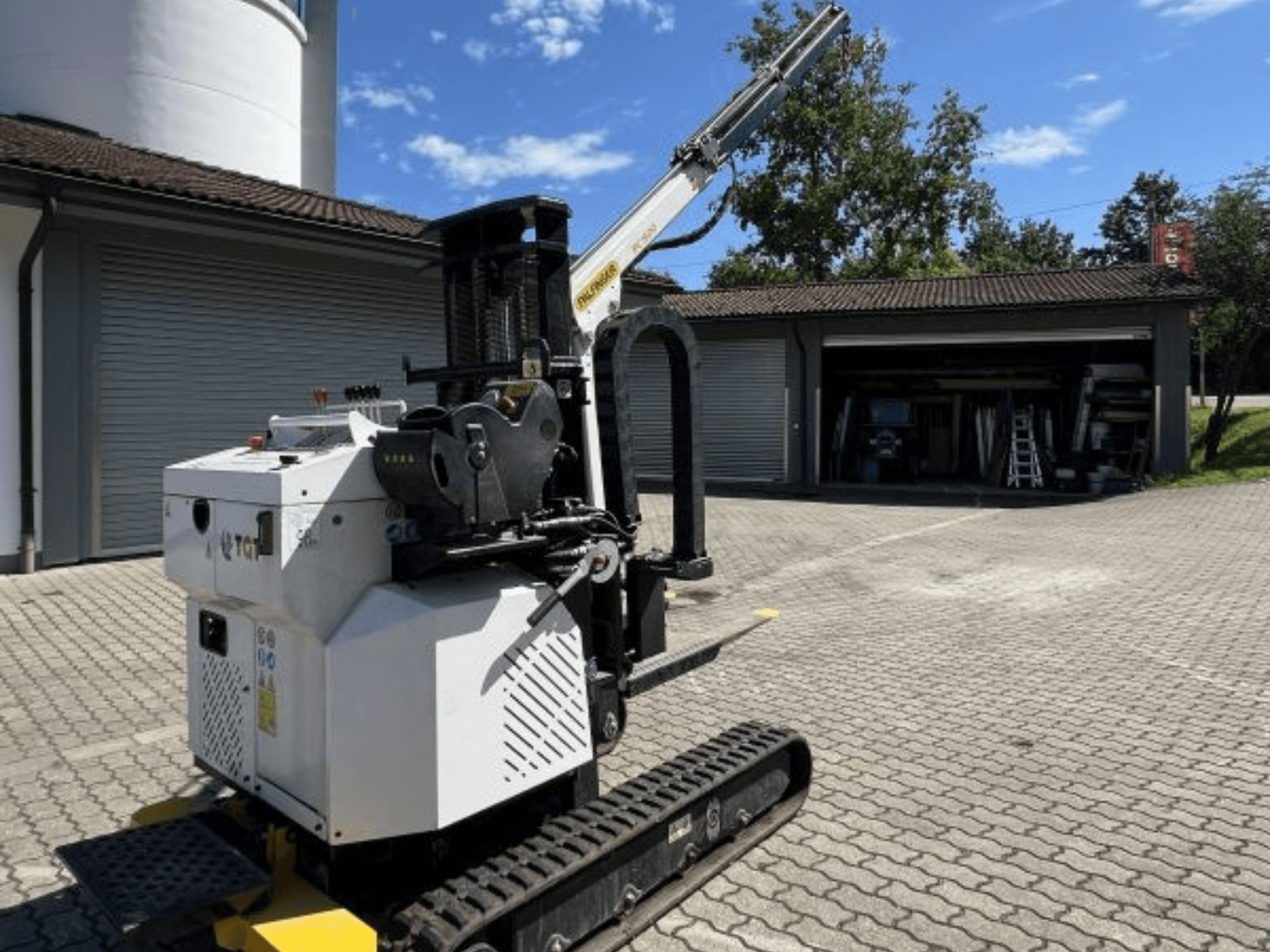 TGI byggkran, minikran, sidovy, visar larvband och monterad bom, parkerad på en tegelsten vid ett garage.