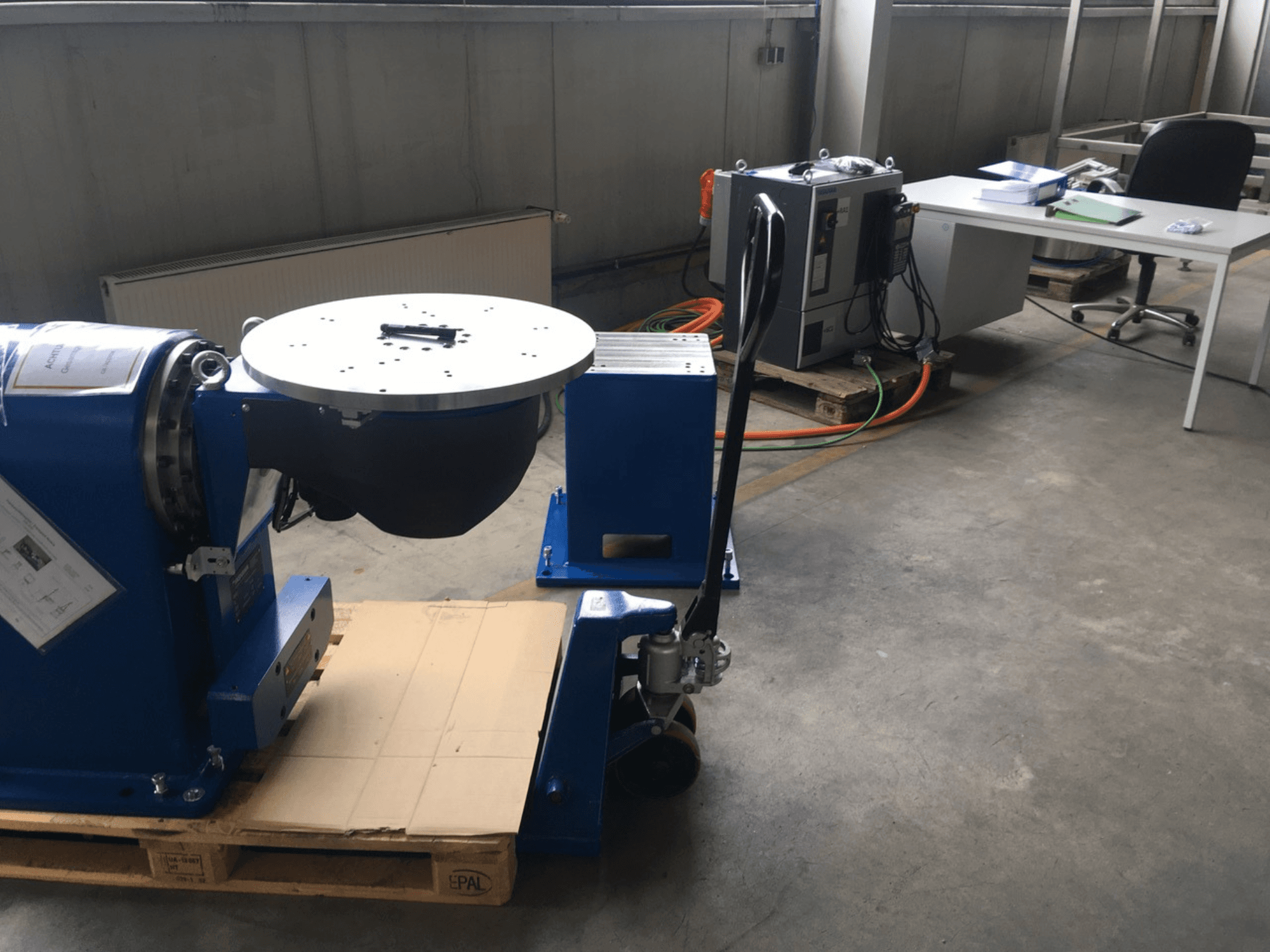 Blå industriutrustning med cirkulär bas och kontrollpanel, sedd framifrån, på en träpall i en kontorsmiljö.