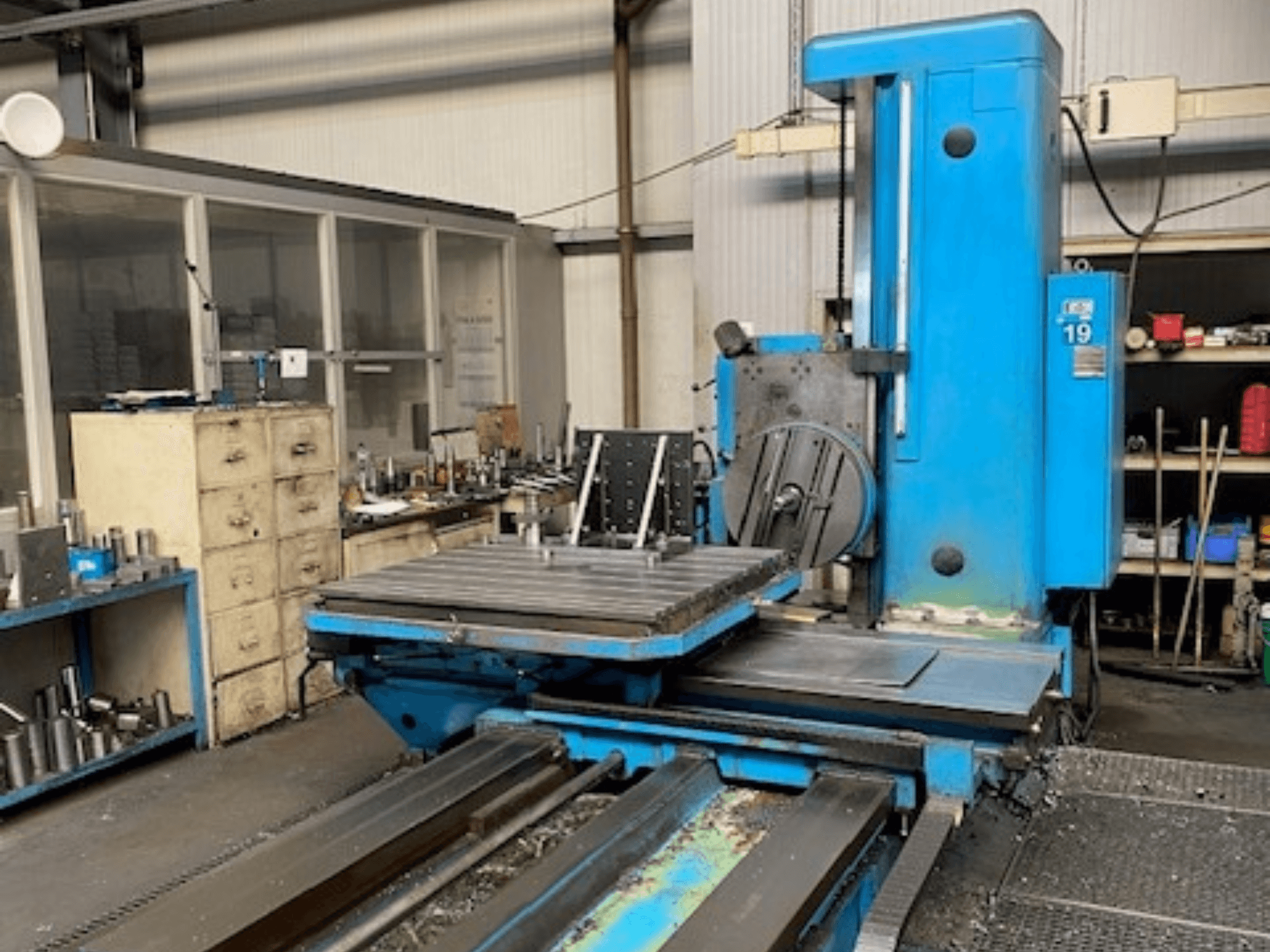 Blå fräsmaskin framifrån, modellnummer 19, på ett verkstadsgolv med verktyg och ett förvaringsskåp i bakgrunden.