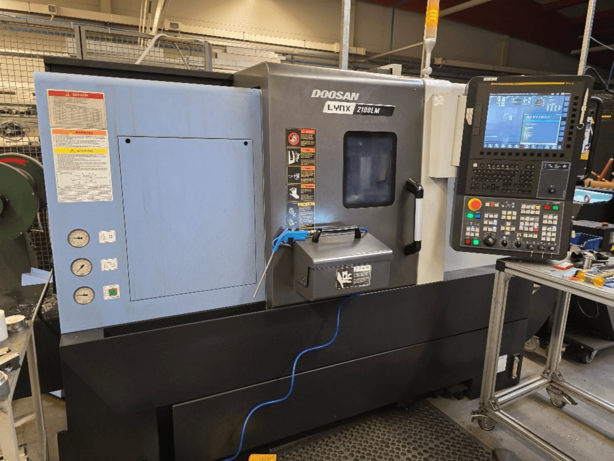 Doosan Lynx 2100LM CNC-svarv, framifrån, med kontrollpanel, tryckmätare och säkerhetsvarningar.