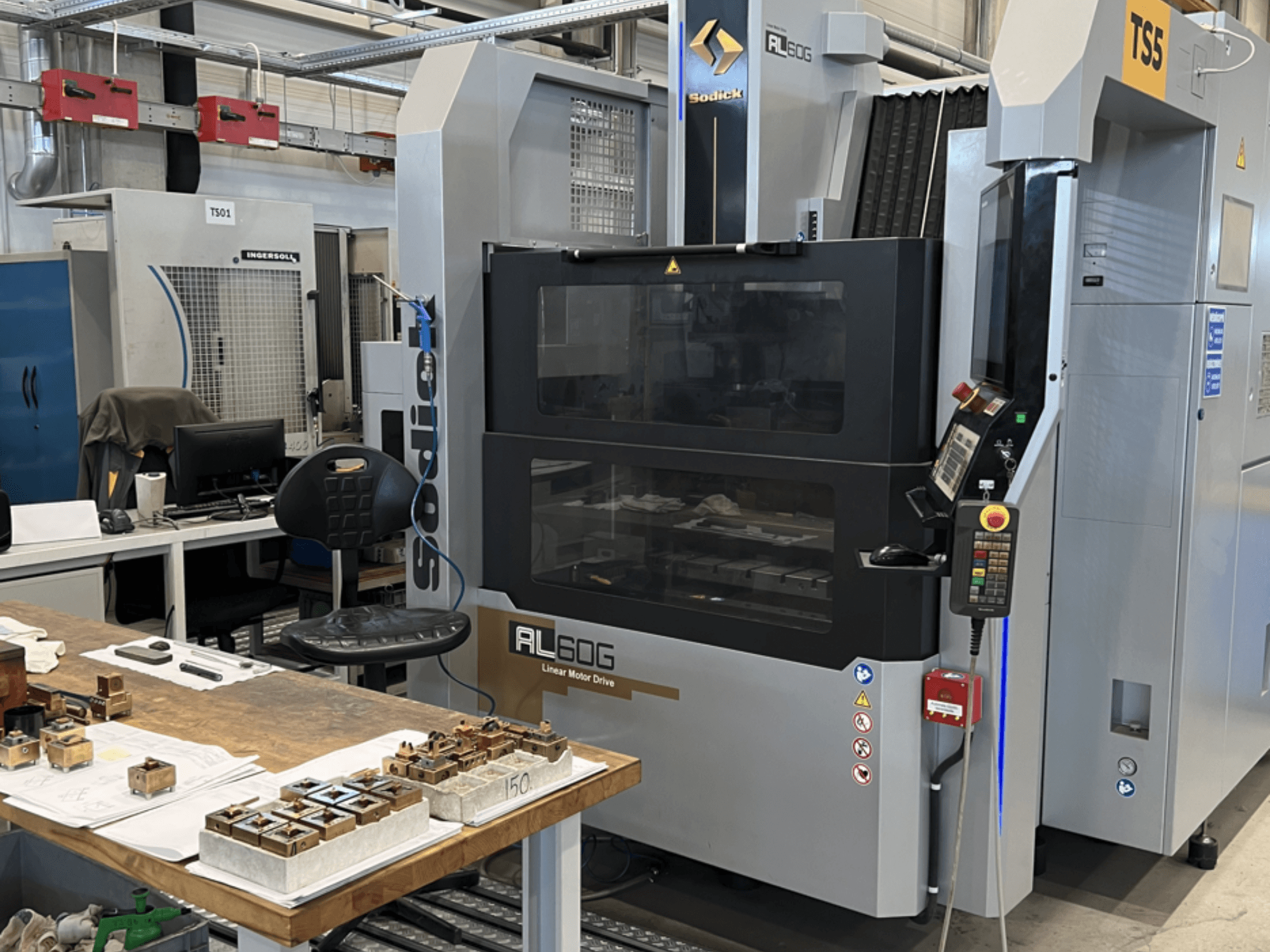 Sodick AL60G Die-Sinking EDM Machine; framifrån visar reglage, fönster och arbetsyta med olika metalldelar.