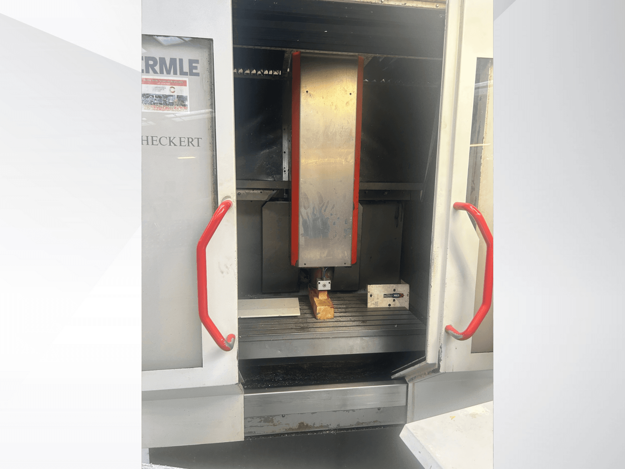 Hermle CNC-maskin framifrån, med metallisk interiör, röda handtag och ett arbetsstycke på bordet.