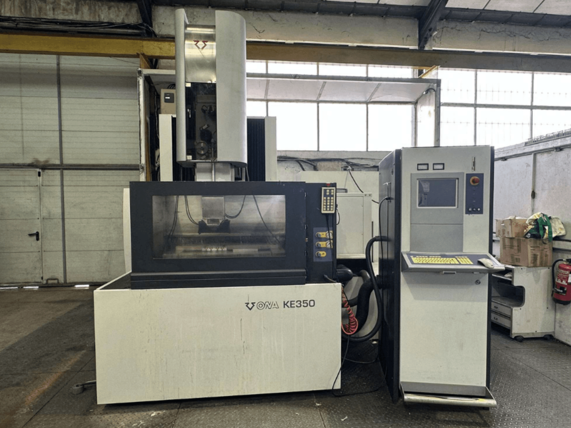 Frontvy av CNC-maskinen ONA KE350 med kontrollpanel, displayskärm och olika knappar, placerad i en industriell miljö.