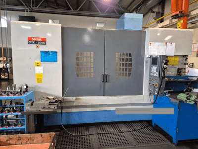 Mazak VTC-300C-maskin i en fabrik, framifrån med kontrollpanel, grå dörrar med fönstergaller och närliggande utrustning.