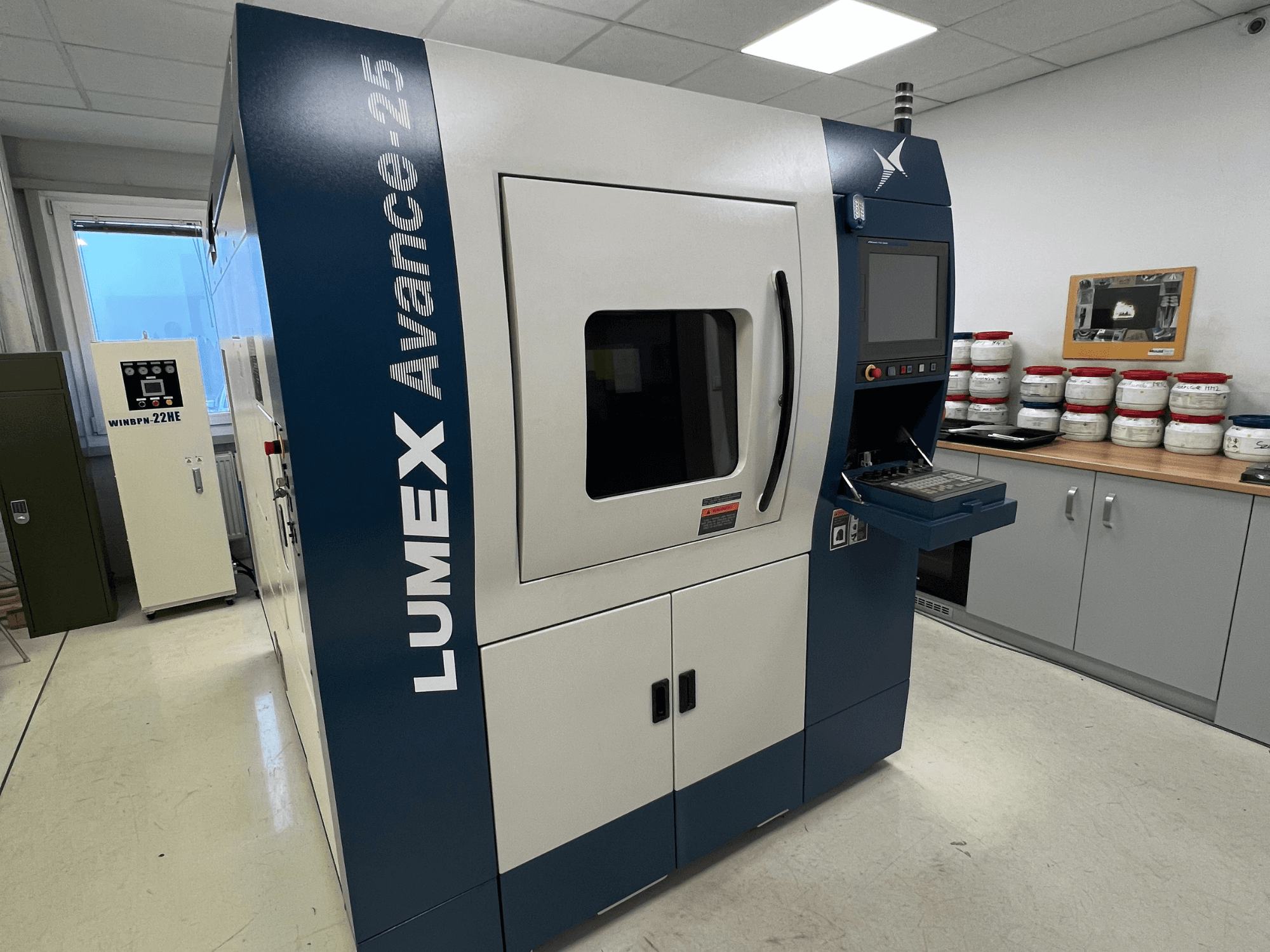 LUMEX Avance-25 3D-skrivare i en labbmiljö, framifrån. Kontrollpanel med skärm och knappar synliga, utrustning i bakgrunden.