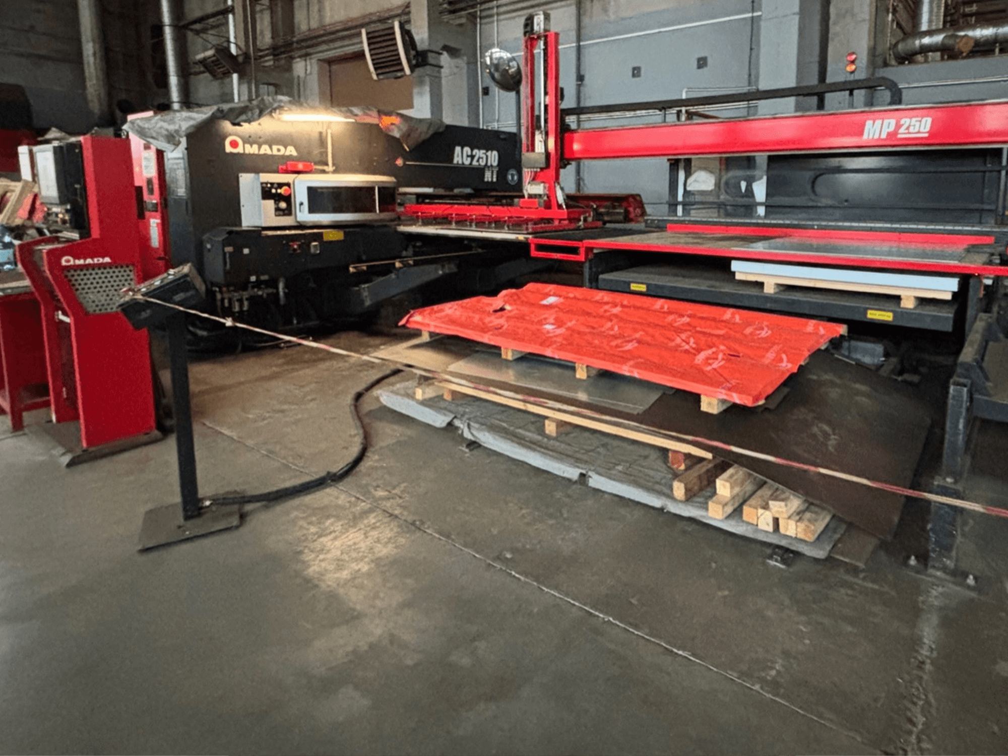 Amada AC2510 NT laserskärare i fabriksmiljö, framifrån, tunga maskiner med metallplåtar och pallar på golvet.