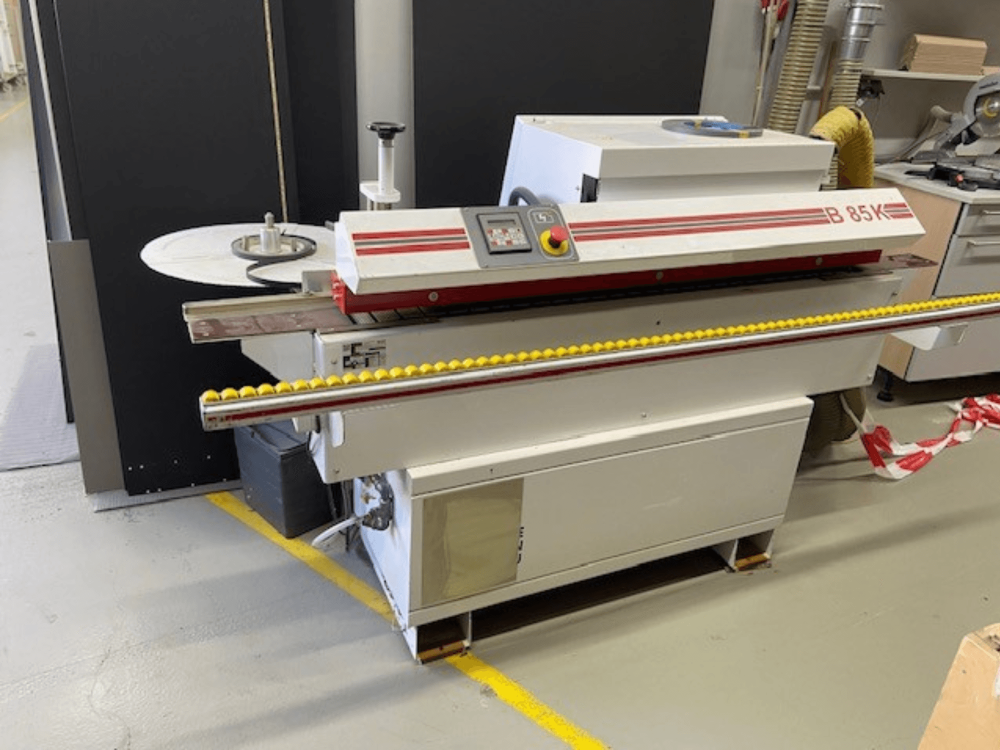 B 85K kantlistningsmaskin i vitt och rött, framifrån, har en kontrollpanel och gul rulle för materialmatning.