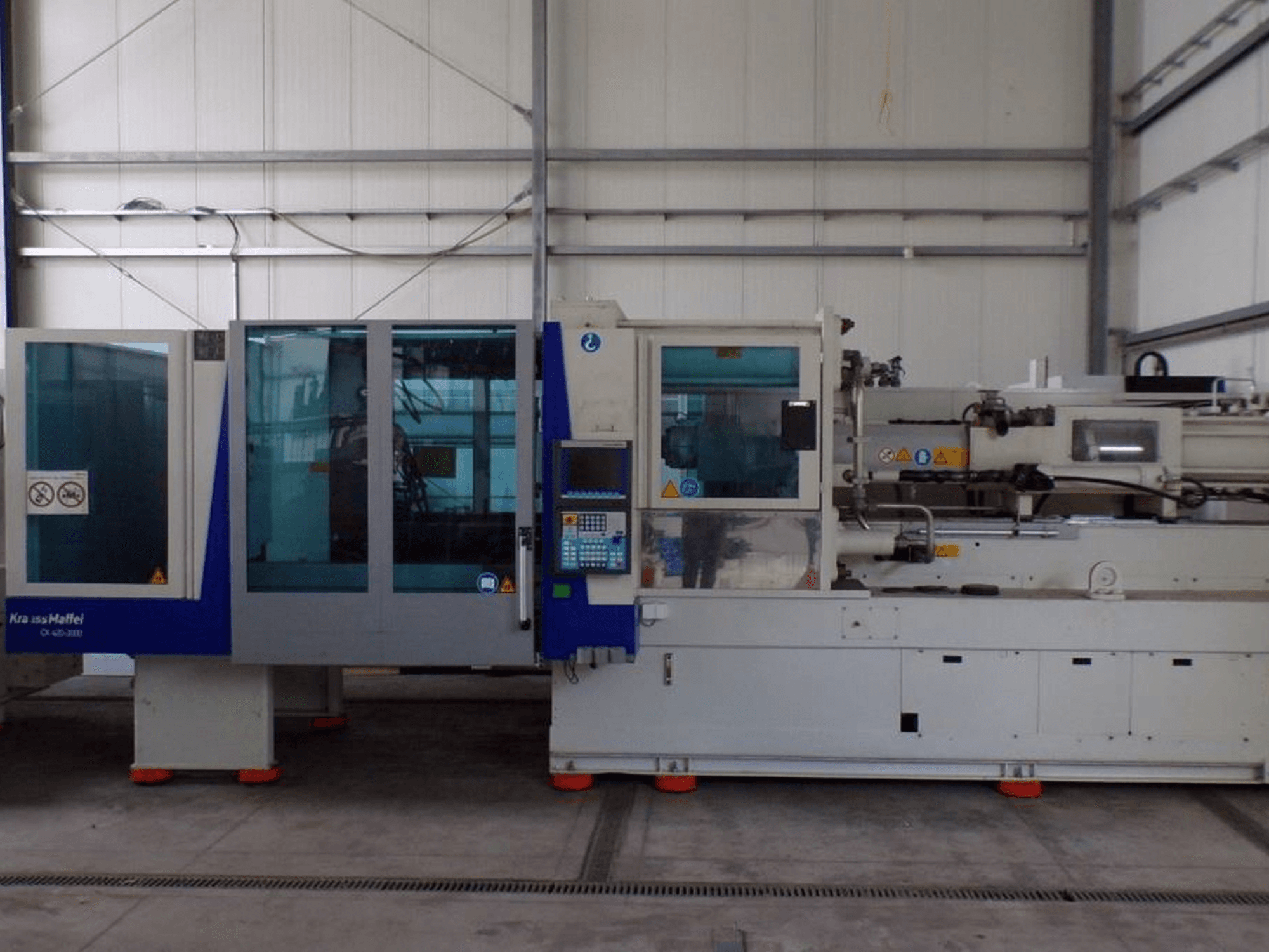 KraussMaffei formsprutningsmaskin, sidovy, med kontrollpanel och säkerhetsindikatorer, synlig i en fabriksmiljö.