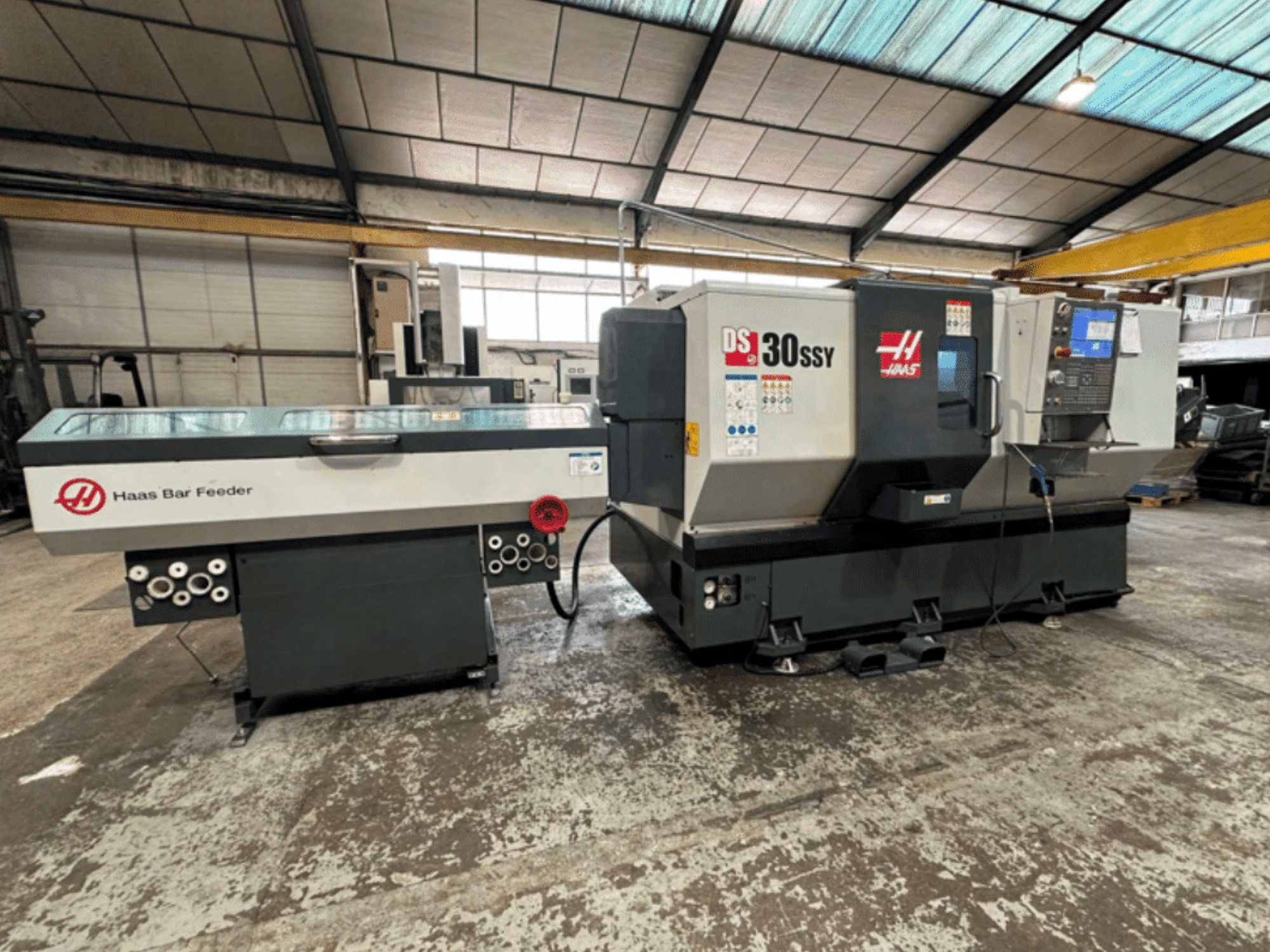 Haas DS-30SSY CNC-svarv med Haas stångmatare, sidovy som visar kontrollpanelen och matningsmekanismen i en verkstadsmiljö.