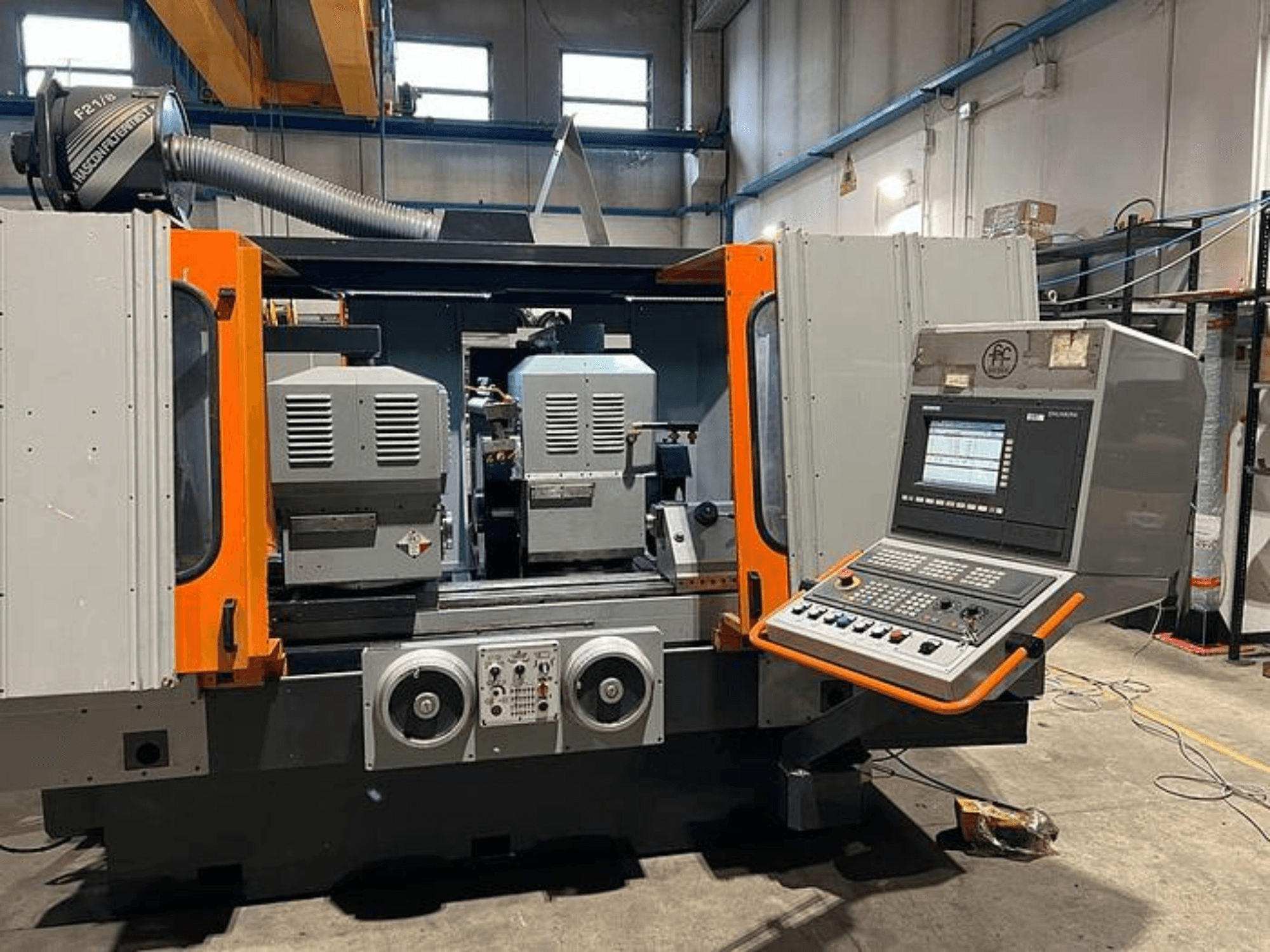 Frontvy av en CNC-maskin med dubbla spindlar, kontrollpanel och orangefärgade detaljer. Med ventilationskanal och manöverreglage.