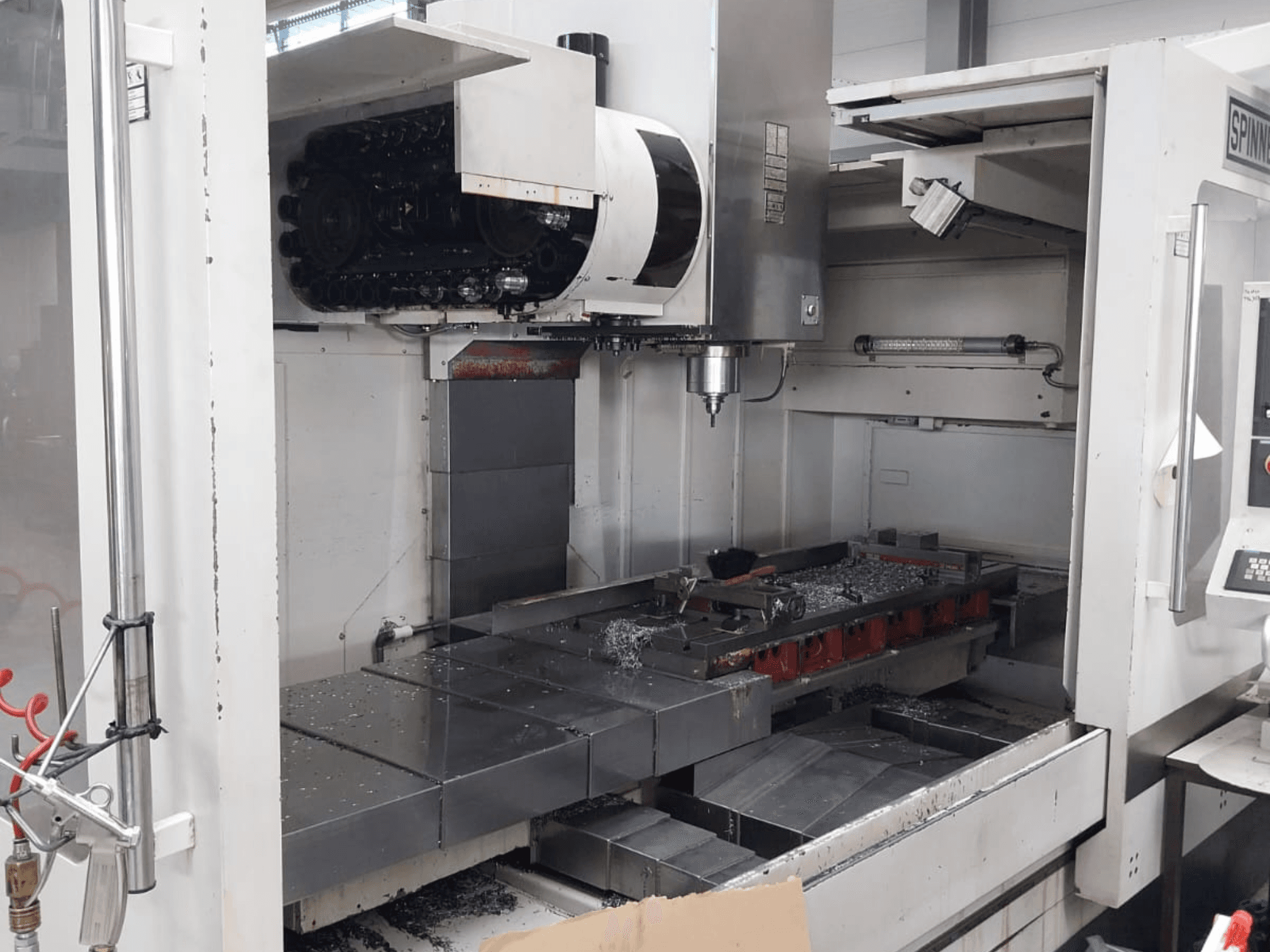 Frontvy av den vertikala fleroperationsmaskinen SPINNER MVC 1600 som visar den öppna arbetsytan, spindeln och metallspånen på bordet.