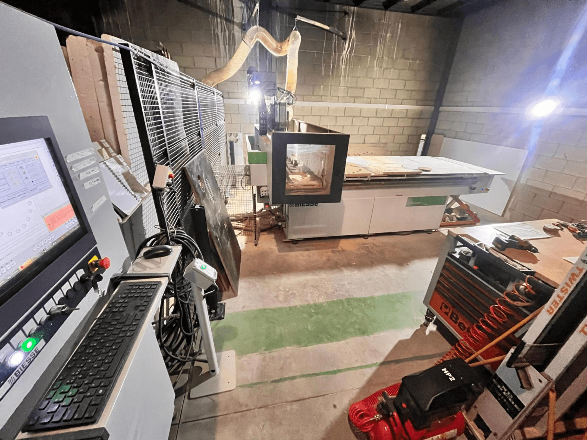 Biesse CNC-maskin i en verkstad, sidovy som visar kontrollpanel, dammutsug, verktyg och olika material på bordet.