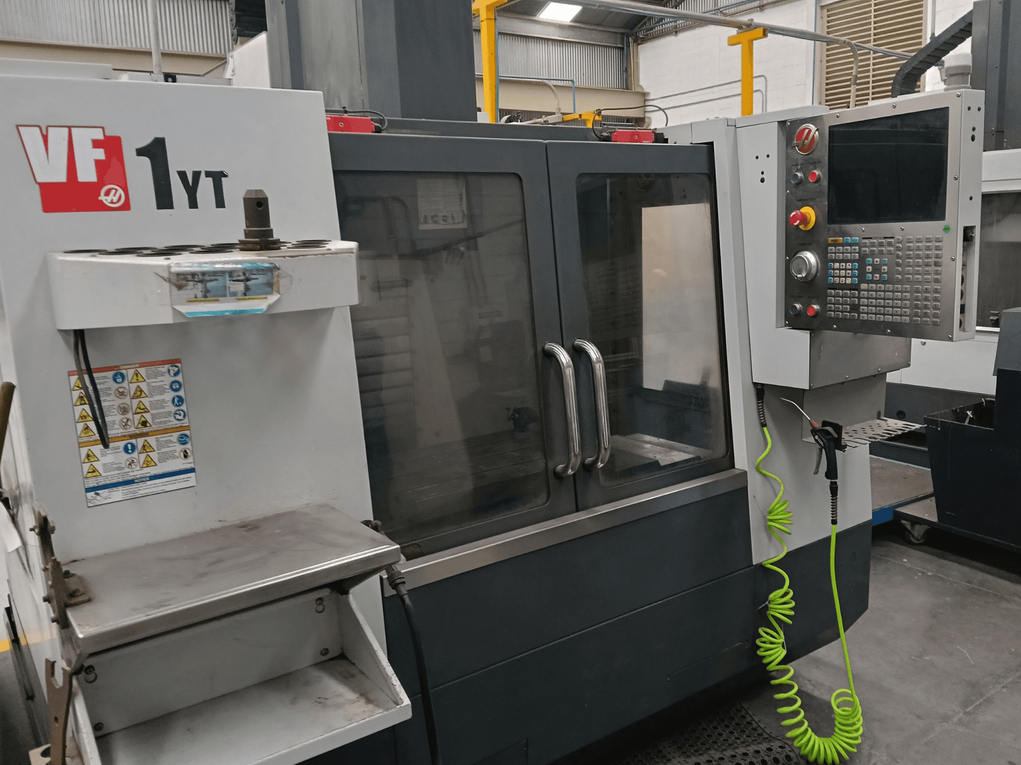 HAAS VF-1YT-2 vertikalt bearbetningscenter, framifrån, med kontrollpanel, verktygshållare, säkerhetsetiketter och bearbetningsområde.