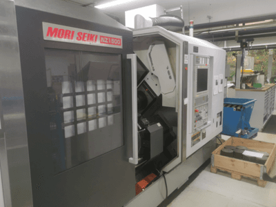 MORI SEIKI NZ1500 CNC-maskin i en liten vinkel, visar kontrollpanelen och arbetsytan med arbetsstycken i metall på en träpall.