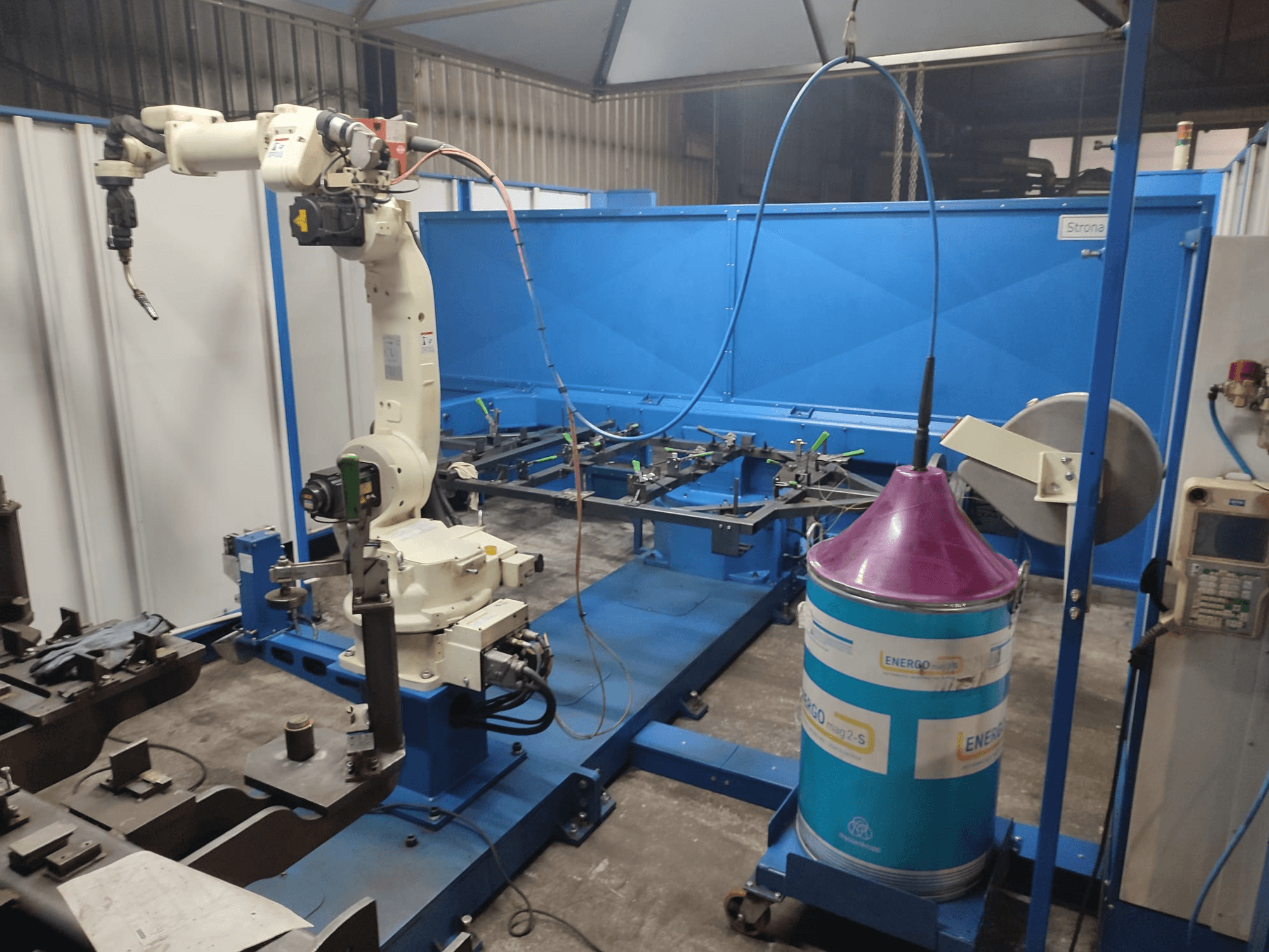 Industriell robotarm med vit och blå bas, uppställd i en verkstad med blå arbetsyta och metallkomponenter.