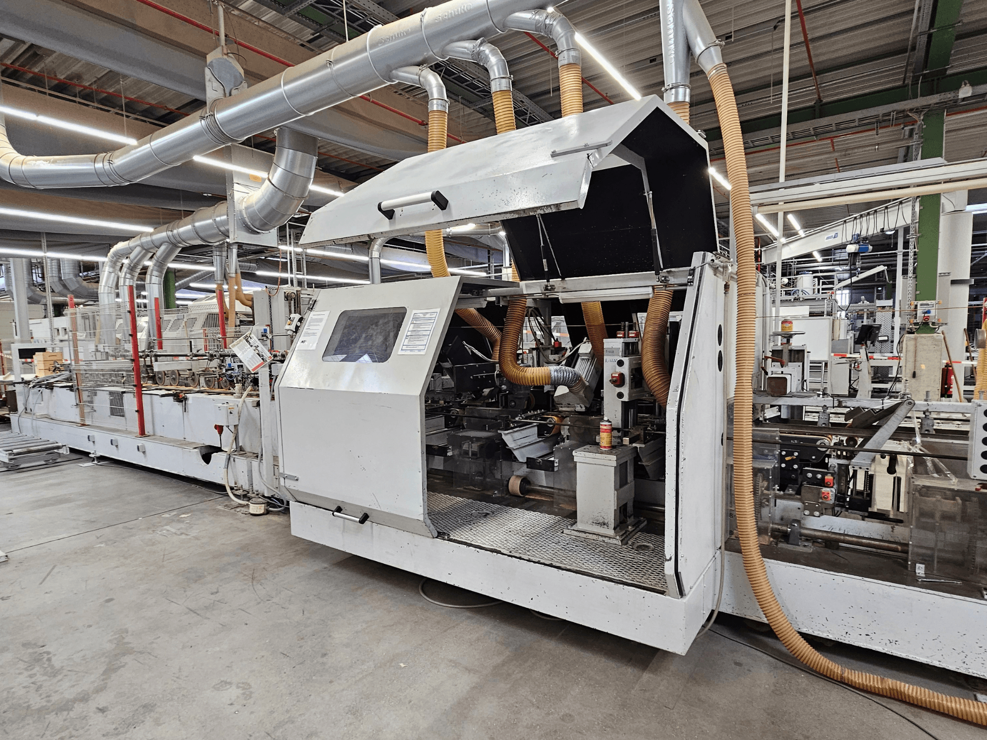 HOMAG FG63 kantlistmaskin i fabriksmiljö, sidovy som visar driftskomponenter och dammutsugningssystem.