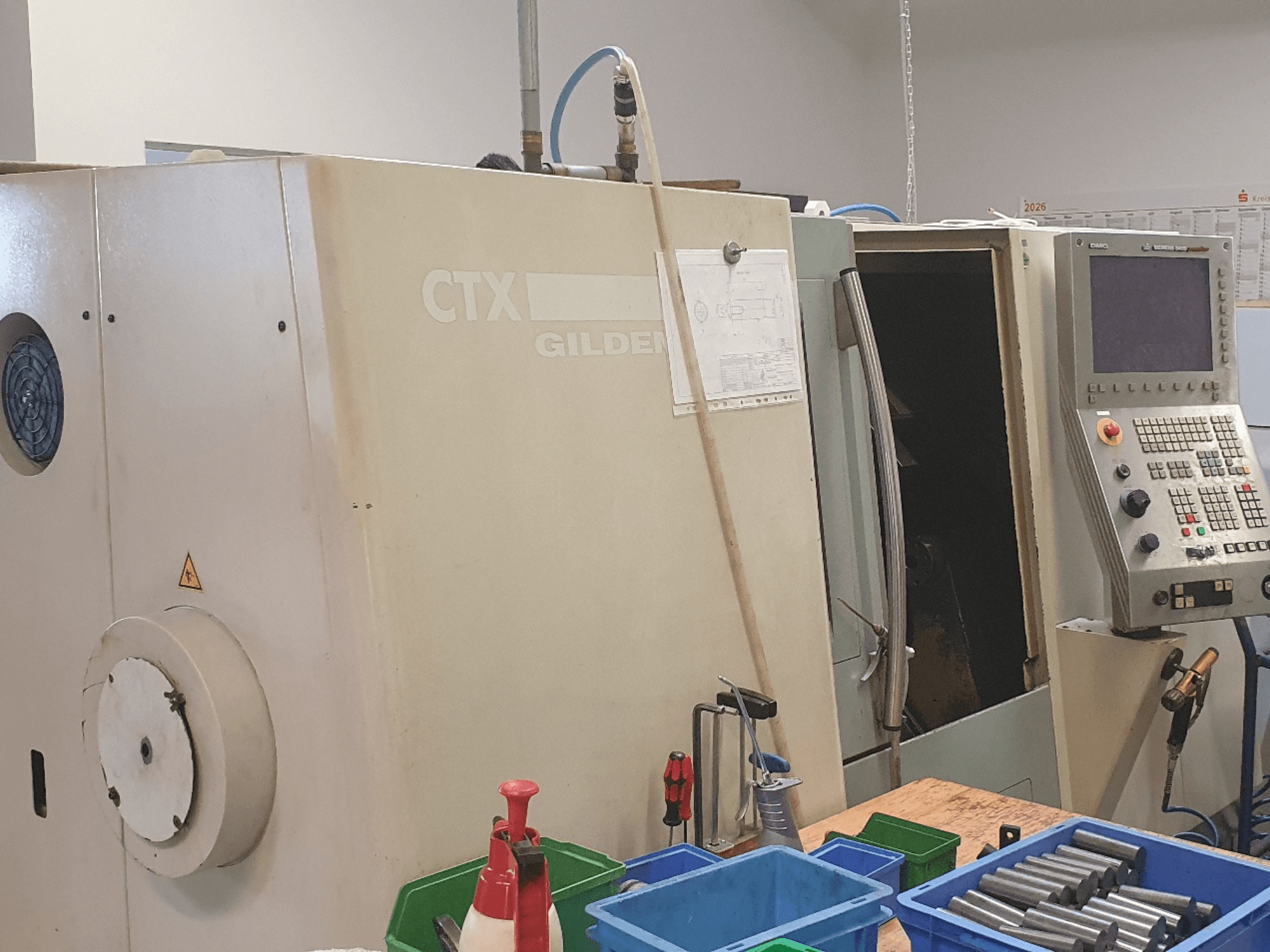Framifrånvy av den horisontella svarven DMG GILDEMEISTER CTX 310, med kontrollpanelen synlig och verktyg samt behållare i förgrunden.