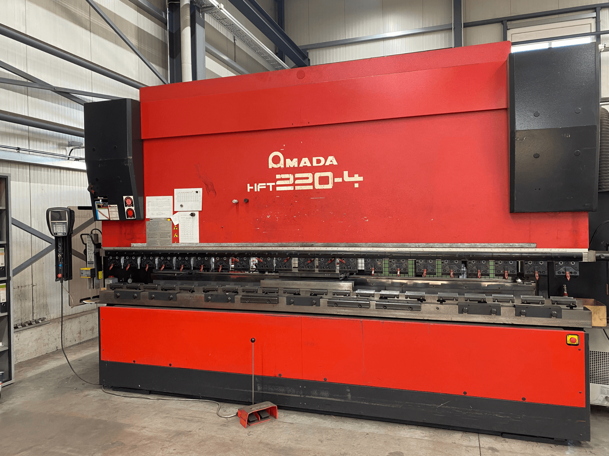 Röd AMADA HFT 220-4 kantpress i fabriksmiljö, framifrån med kontrollpanel och bockverktyg på arbetsytan.
