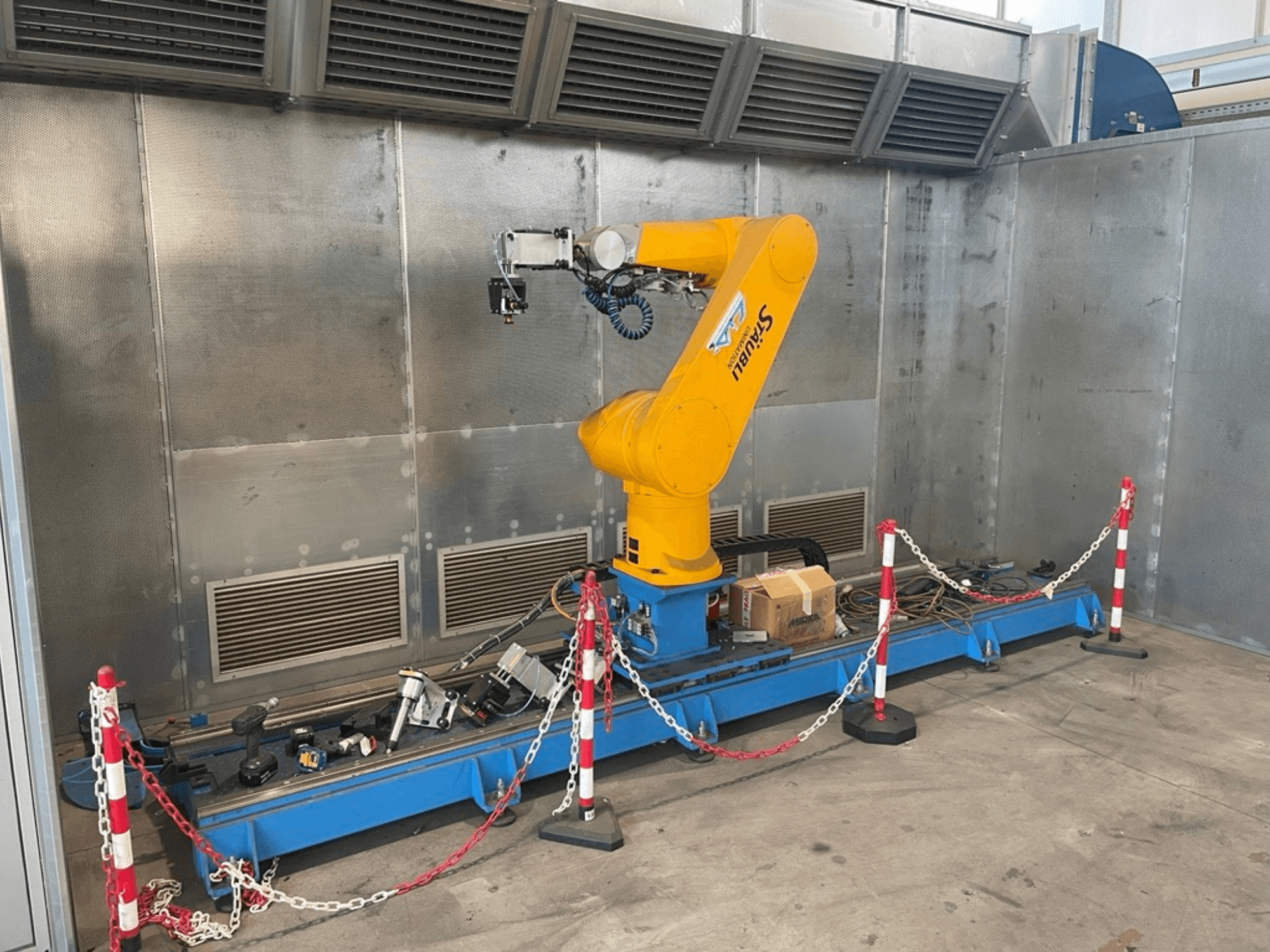 STAUBLI CS8C-RX160 robotarm i en verkstad, vy från sidan, med kablar och olika verktyg på en blå bas omgiven av skyddsbarriärer.