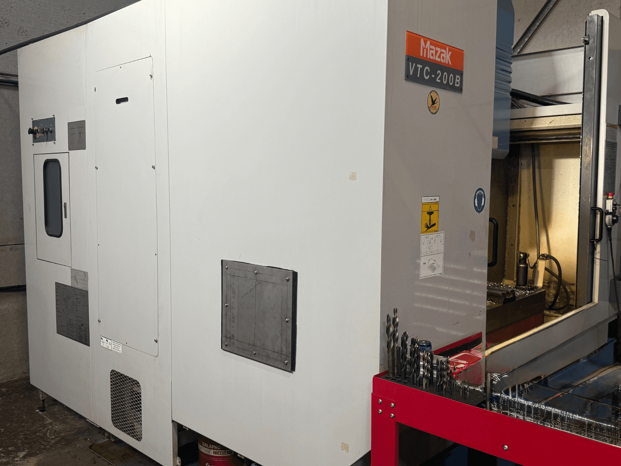 Mazak VTC-200B vertikalt bearbetningscenter sett från sidan, med märkta reglage och borr på ett rött förvaringsställ.