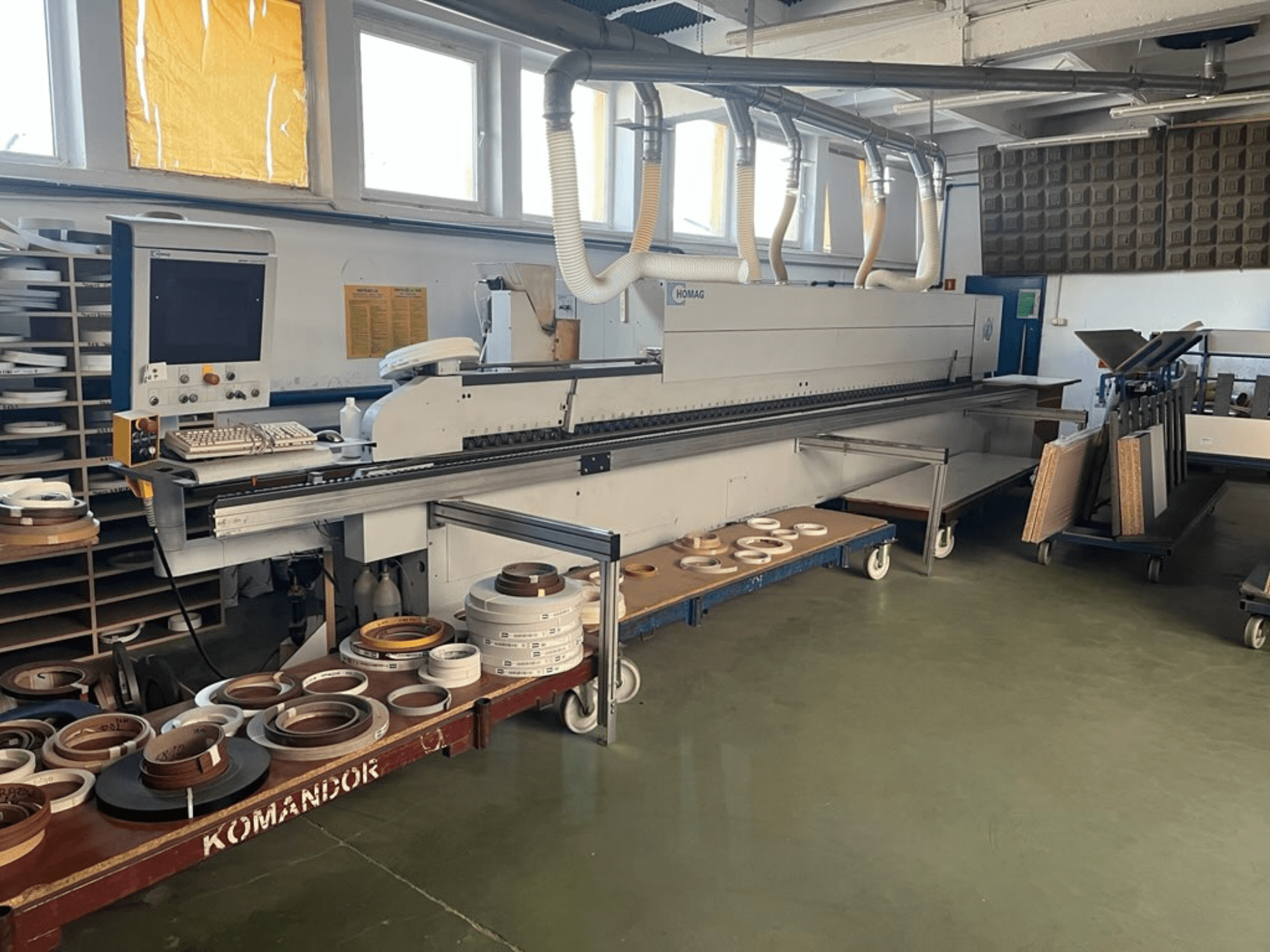 HOMAG kantlistningsmaskin i en verkstad, med reglage och olika kantlistningsmaterial på en blå vagn. Vy från sidan.
