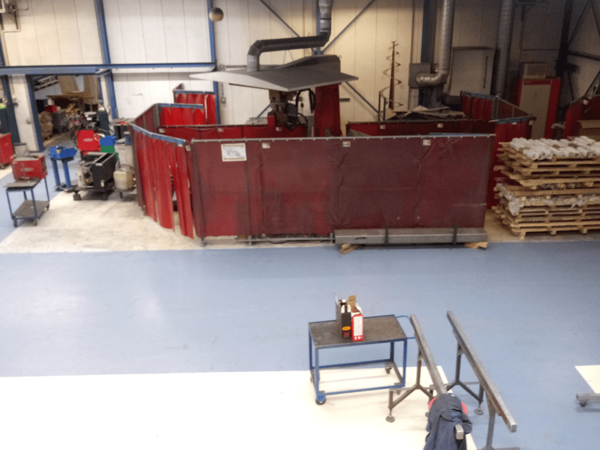 Industriellt arbetsutrymme med röda svetsgardiner, maskiner och pallar; vy från ovan som visar layout och utrustning.