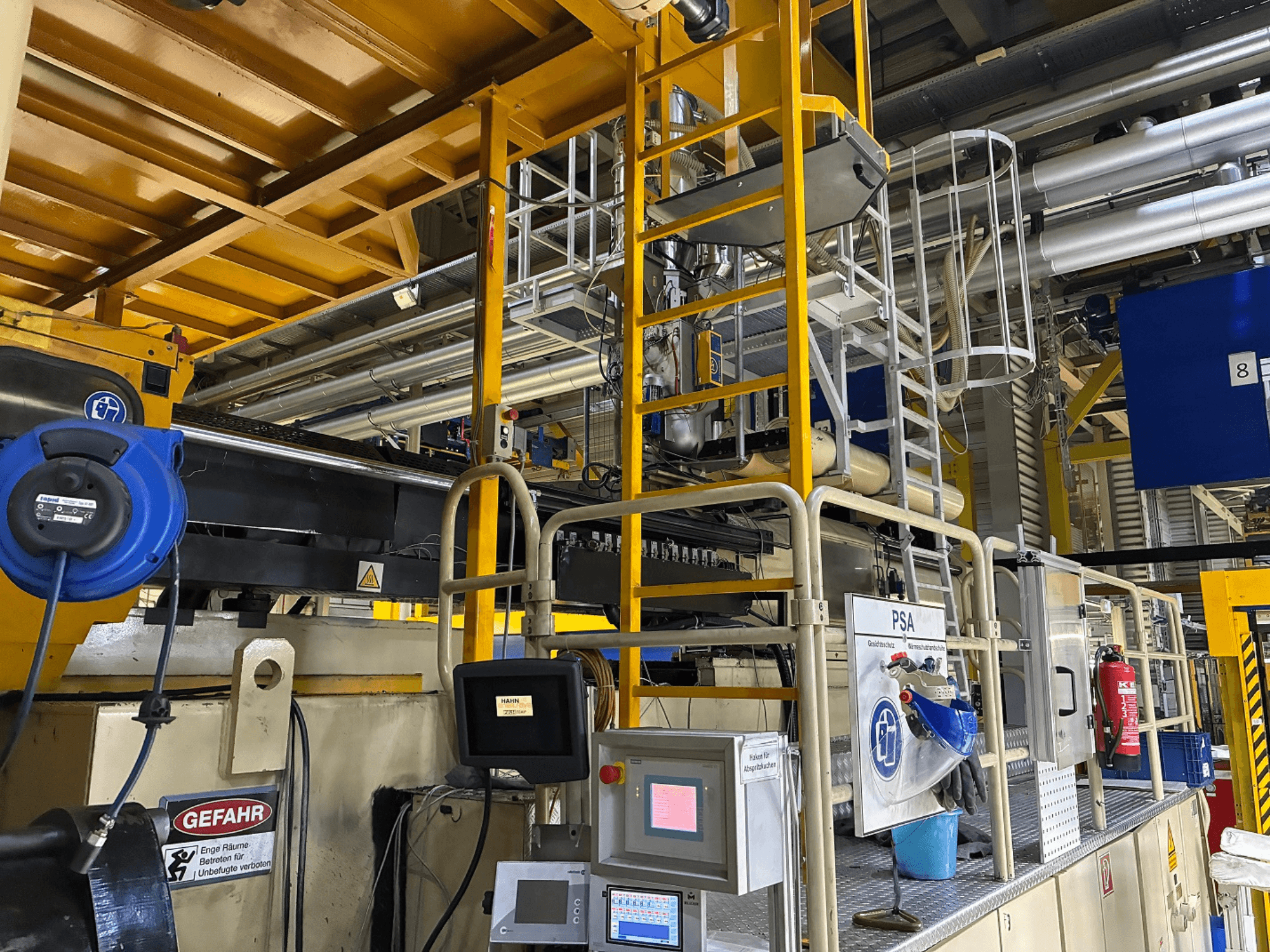 Kontrollpanel för industrimaskiner med blå säkerhetsutrustning och gul byggnadsställning, sidovy som visar rör och varningsskyltar.