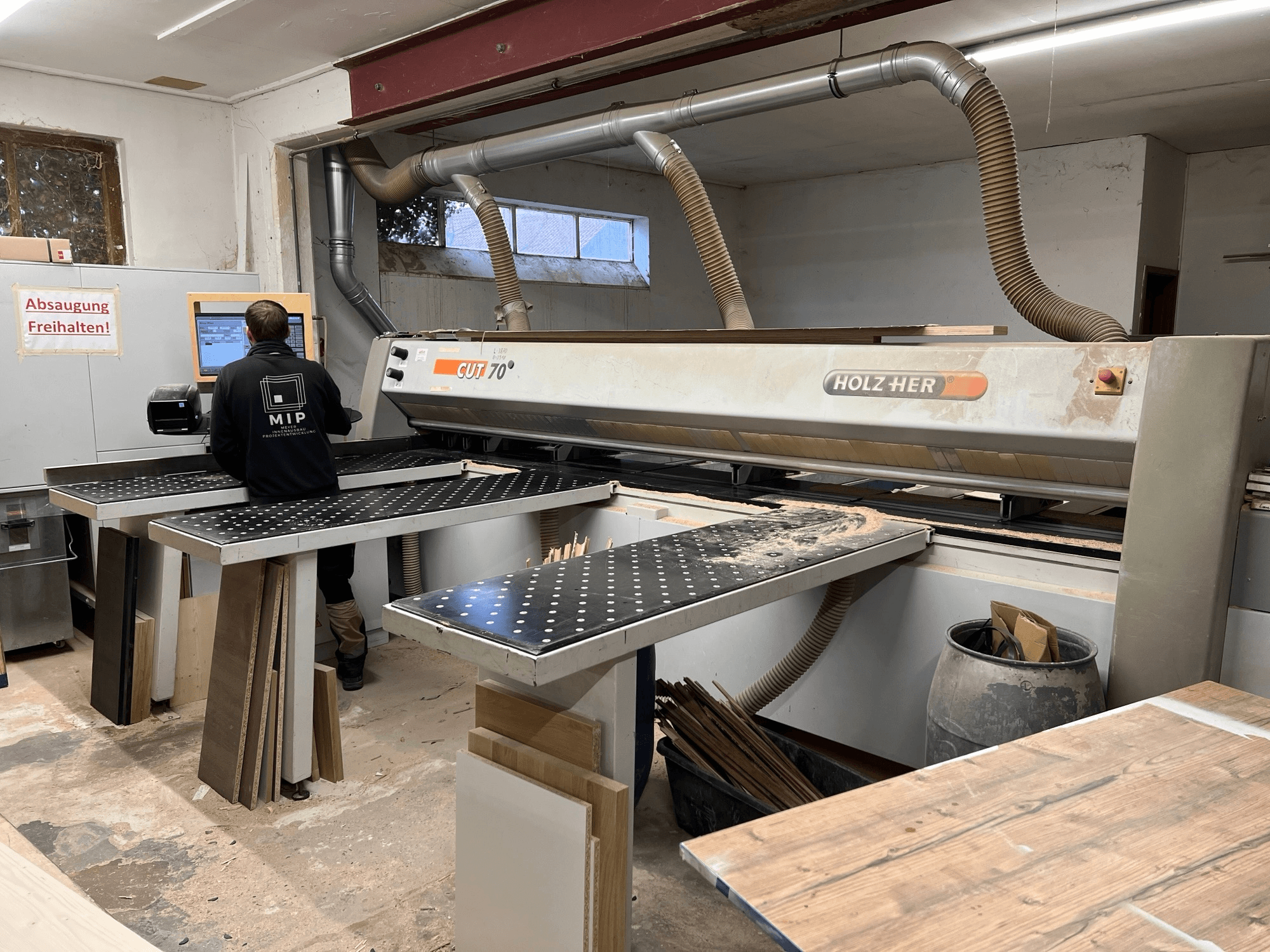 HOLZHER CUT 70 skärmaskin i verkstad; vy framifrån som visar operatör, arbetsbord och trävaror staplade på ytor.